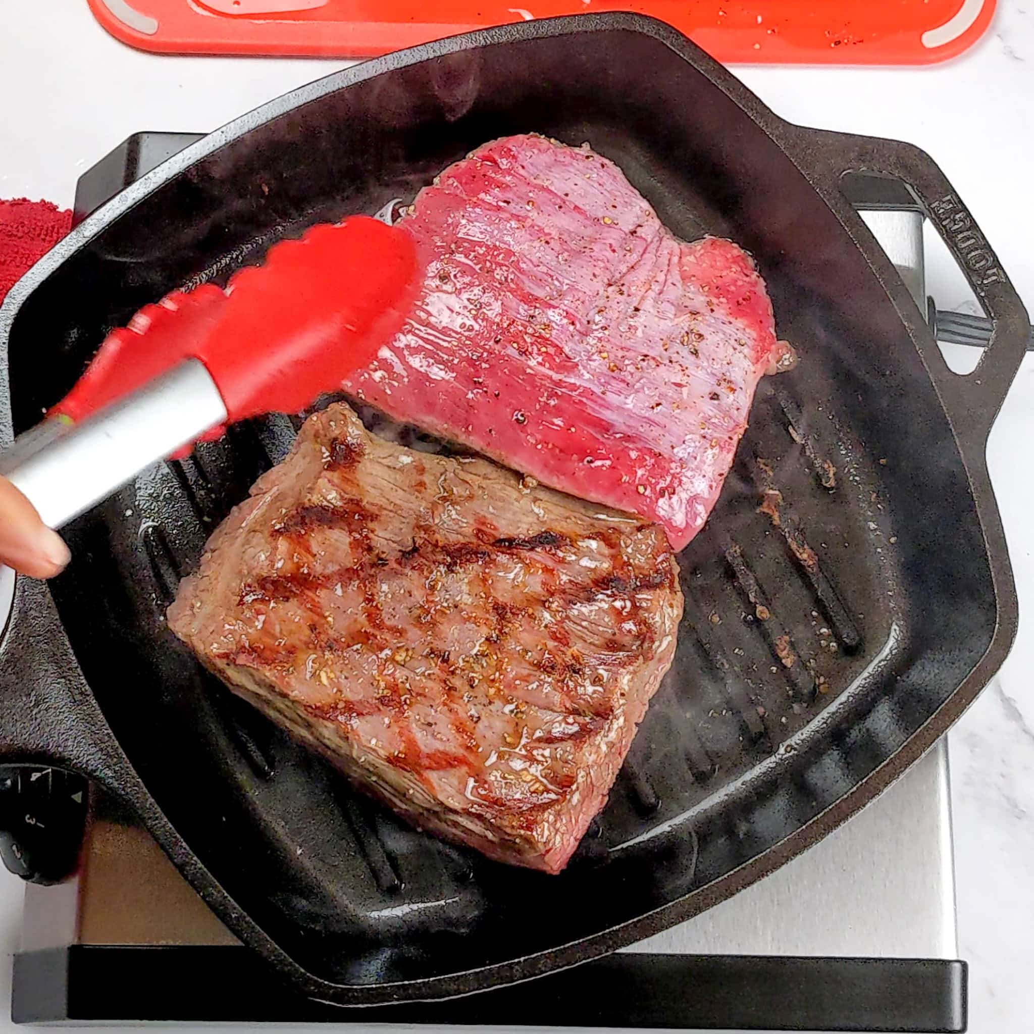 raw portioned and seasoned flank steak in a grill pan with one of them flipped showing a diamond grill mark pattern.