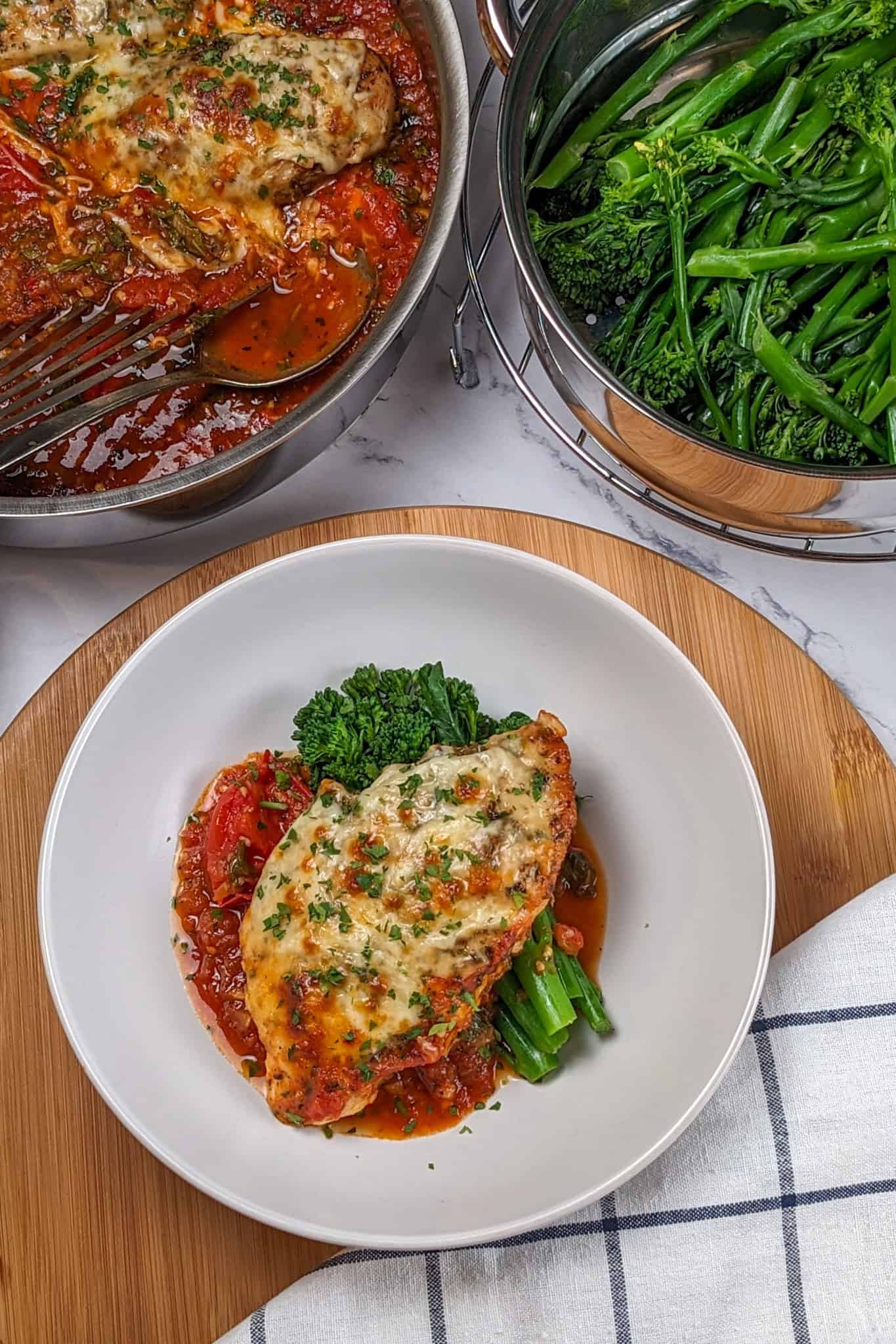 Baked Cheesy Spicy Tomato Basil Chicken and Broccolini on a wide rim bowl surrounded by a saute pan with more baked chicken breast topped with melted mozzarella and steamed broccolini.