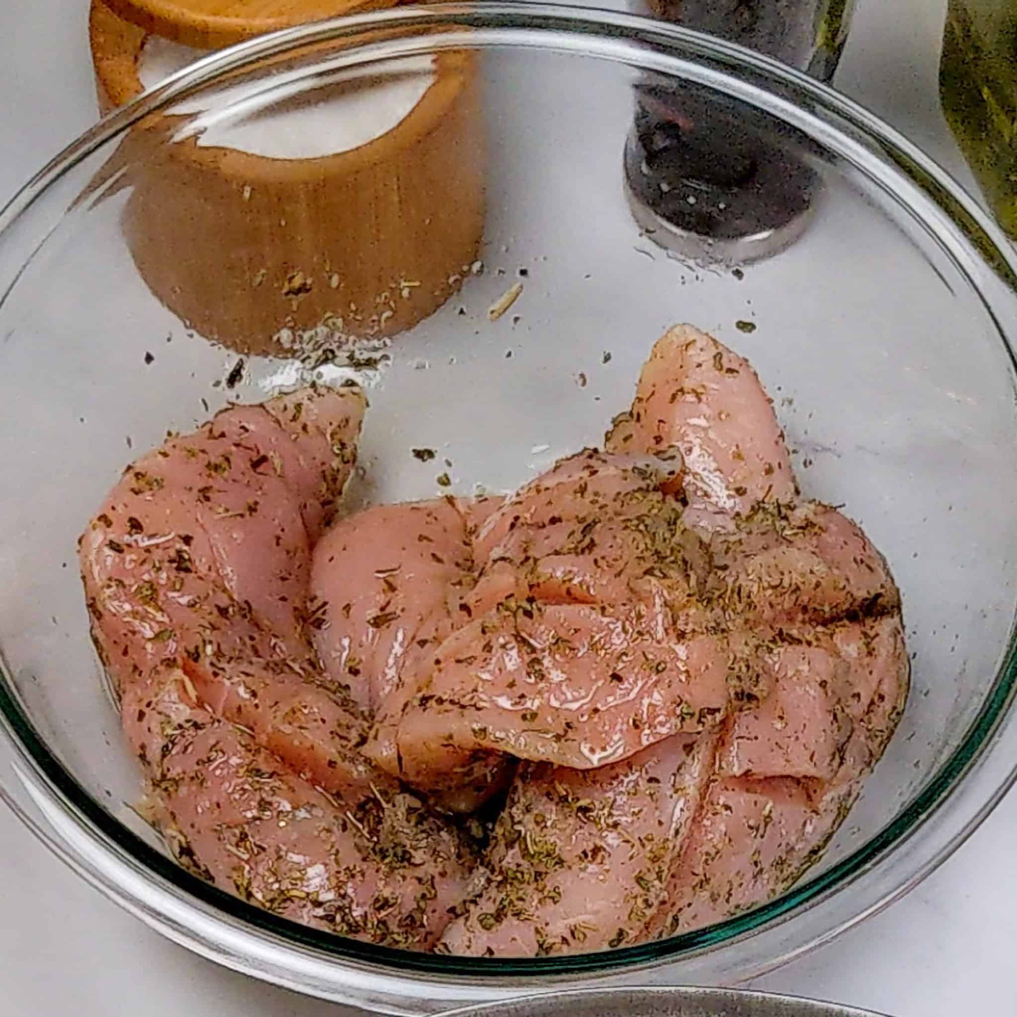 raw diamond slit chicken breast seasoned with Italian herb seasoning and olive oil in a glass mixing bowl.