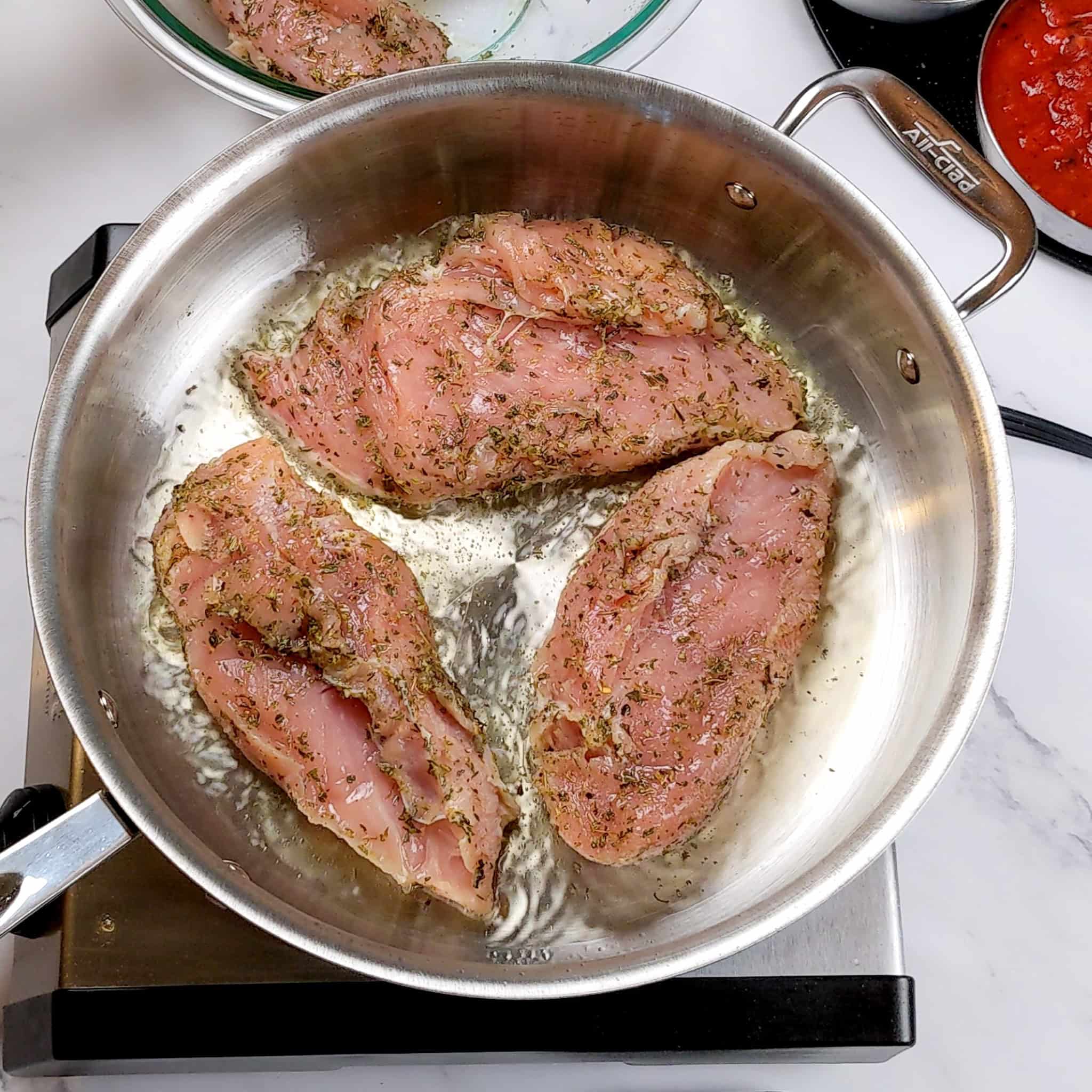 Italian herb seasoned chicken breast being seared skin side down in a large all-clad saute pan.