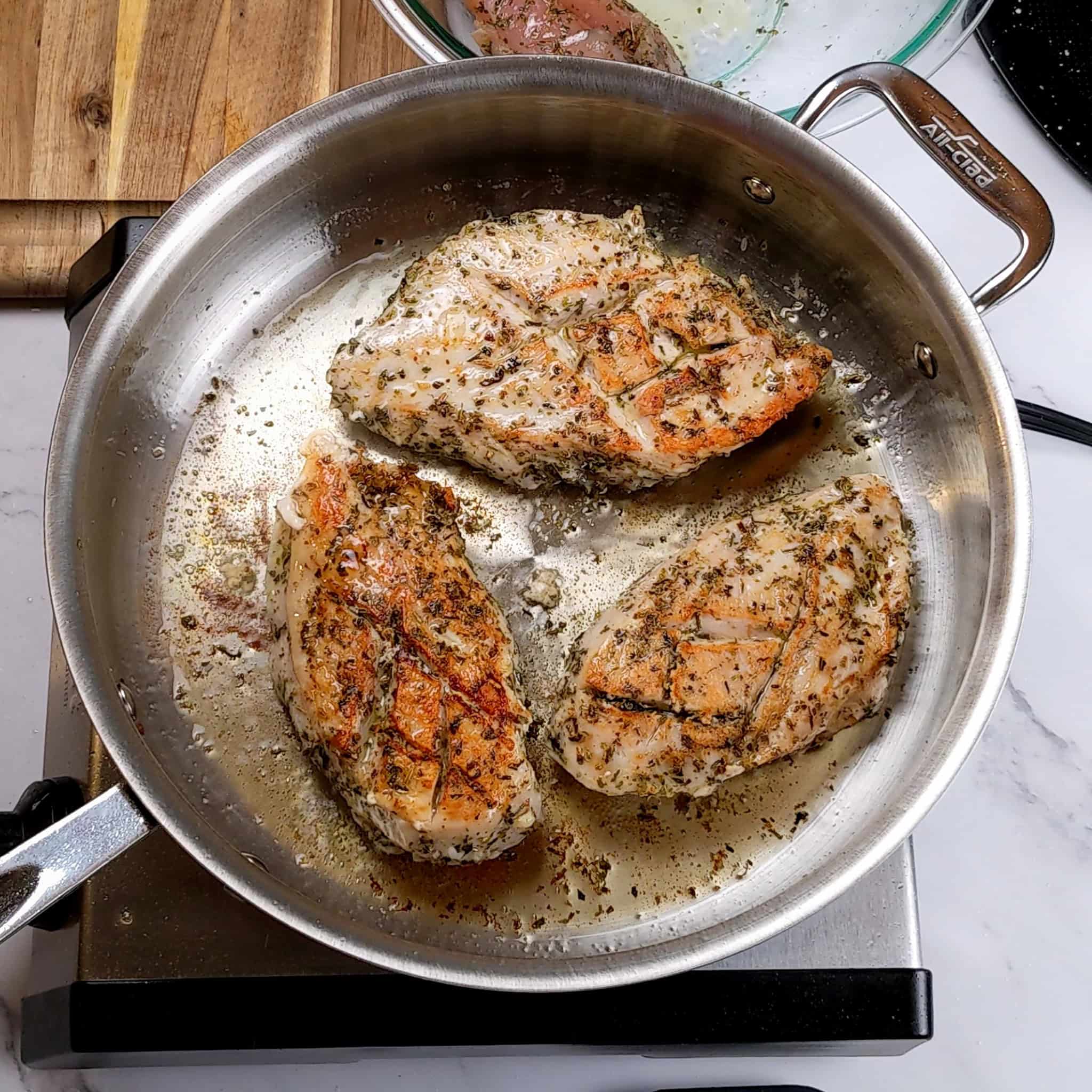 Italian herb seasoned chicken breast being seared skin side up in a large all-clad saute pan.