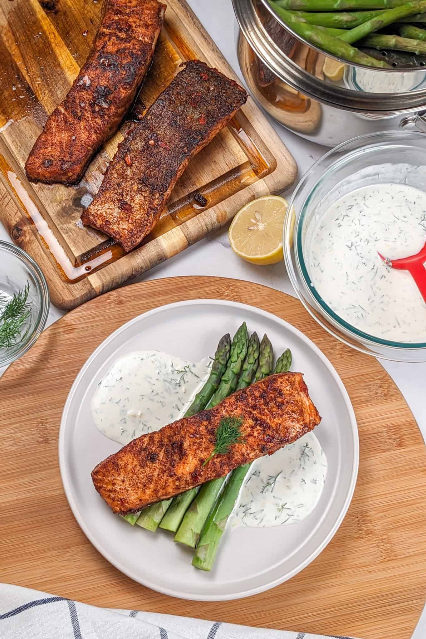 smoked paprika Calabrian Pepper marinated pan-seared Salmon with Creamy Lemon Dill Sauce and jumbo asparagus on a round white plate. Surrounded by more salmon on a cutting board, asparagus and the lemon dill sauce.