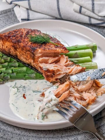 smoked paprika Calabrian Pepper marinated pan-seared Salmon with Creamy Lemon Dill Sauce and jumbo asparagus on a round white plate being eaten with a fork and knife.