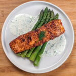 smoked paprika Calabrian Pepper marinated pan-seared Salmon with Creamy Lemon Dill Sauce and jumbo asparagus on a round white plate.
