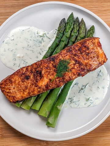 smoked paprika Calabrian Pepper marinated pan-seared Salmon with Creamy Lemon Dill Sauce and jumbo asparagus on a round white plate.