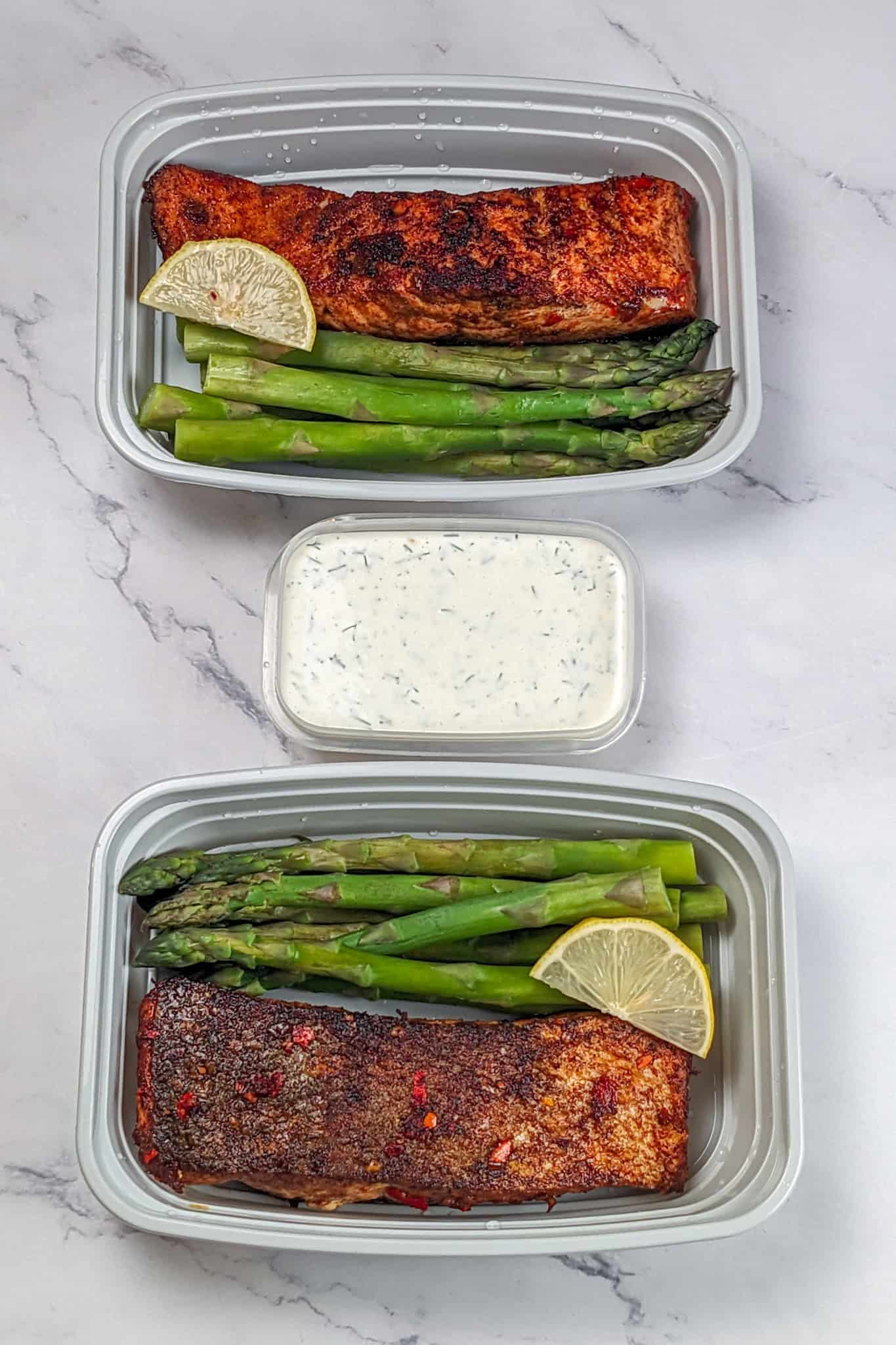 smoke paprika calabrian chili pepper marinated pan seared salmon in a meal prep container wtih jumbo steamed asparagus. with antoher container on the side of the lemon-dill cream sauce.