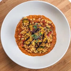 grilled marinated tofu on top of a warm roasted vegetable couscous salad on spicy basil romesco sauce in a wide rim bowl topped with micro greens.