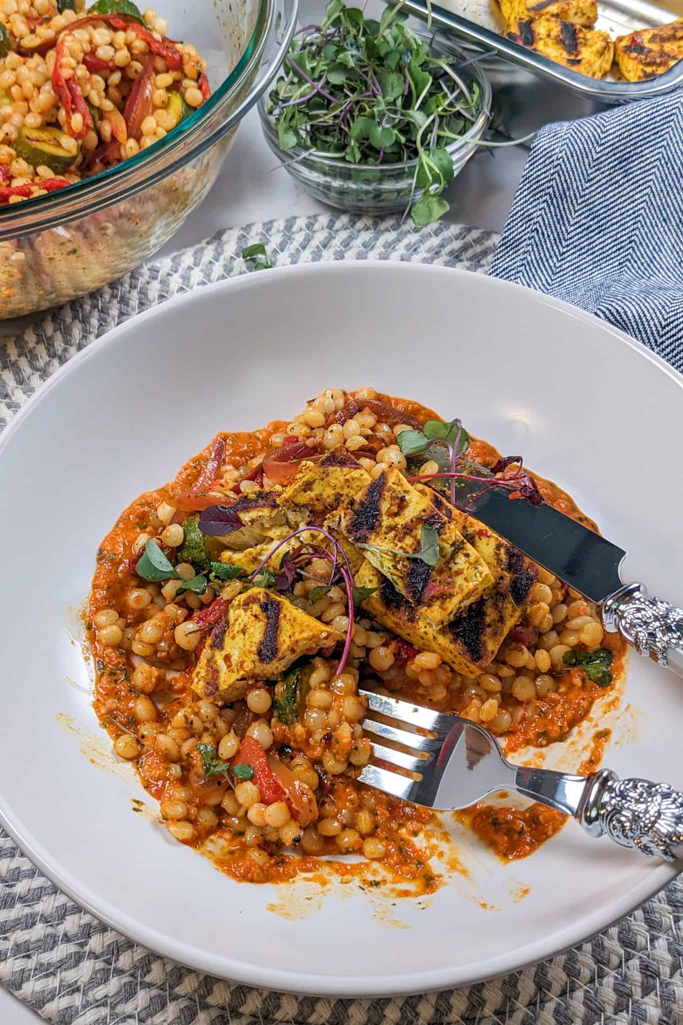 grilled marinated tofu on top of a warm roasted vegetable pearl couscous salad on spicy basil romesco sauce in a wide rim bowl topped with micro greens being eaten with a fork and knife.