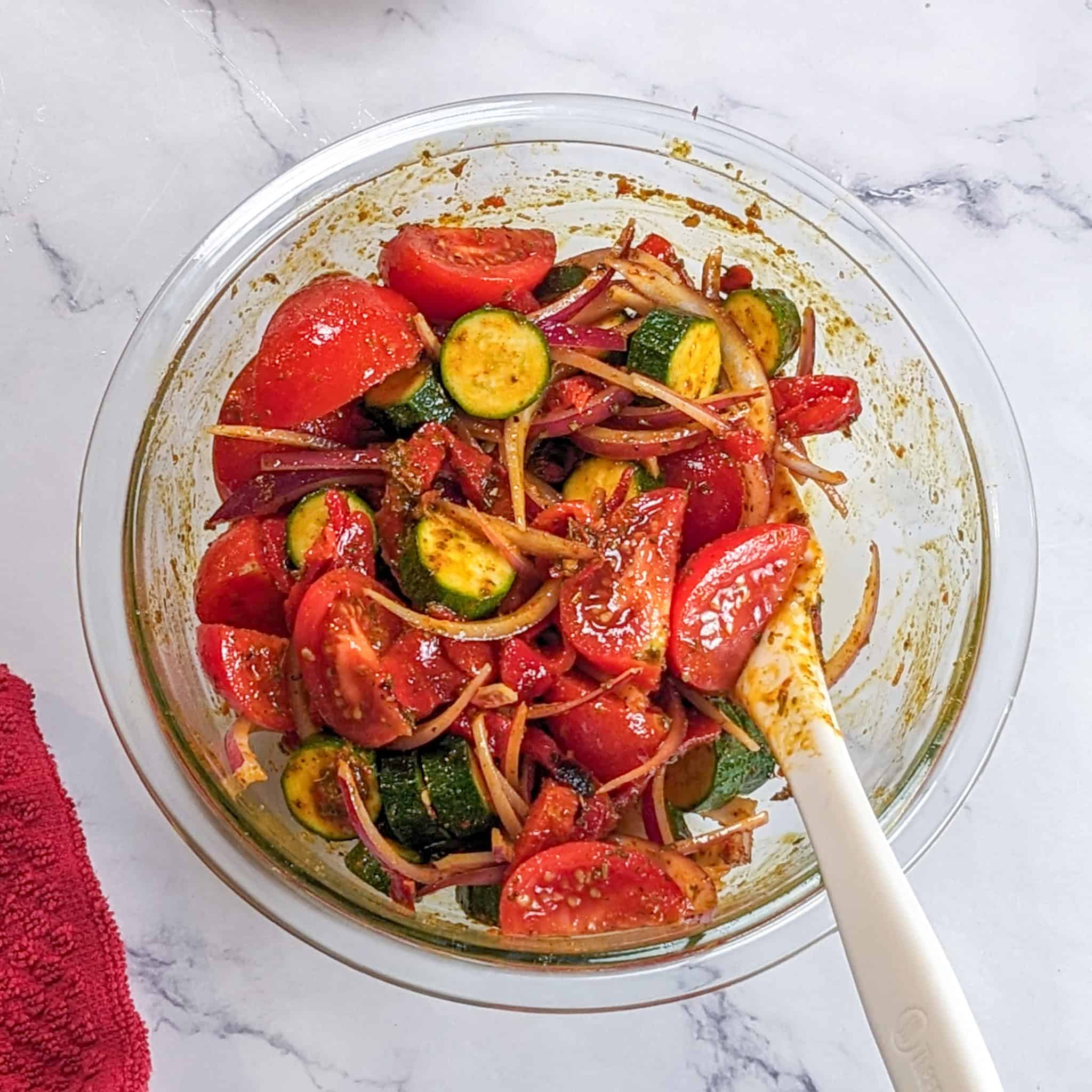 zucchijni, tomatoes, onions, and fire roasted red bell pepper combined with marinade in a glass mixing bowl.