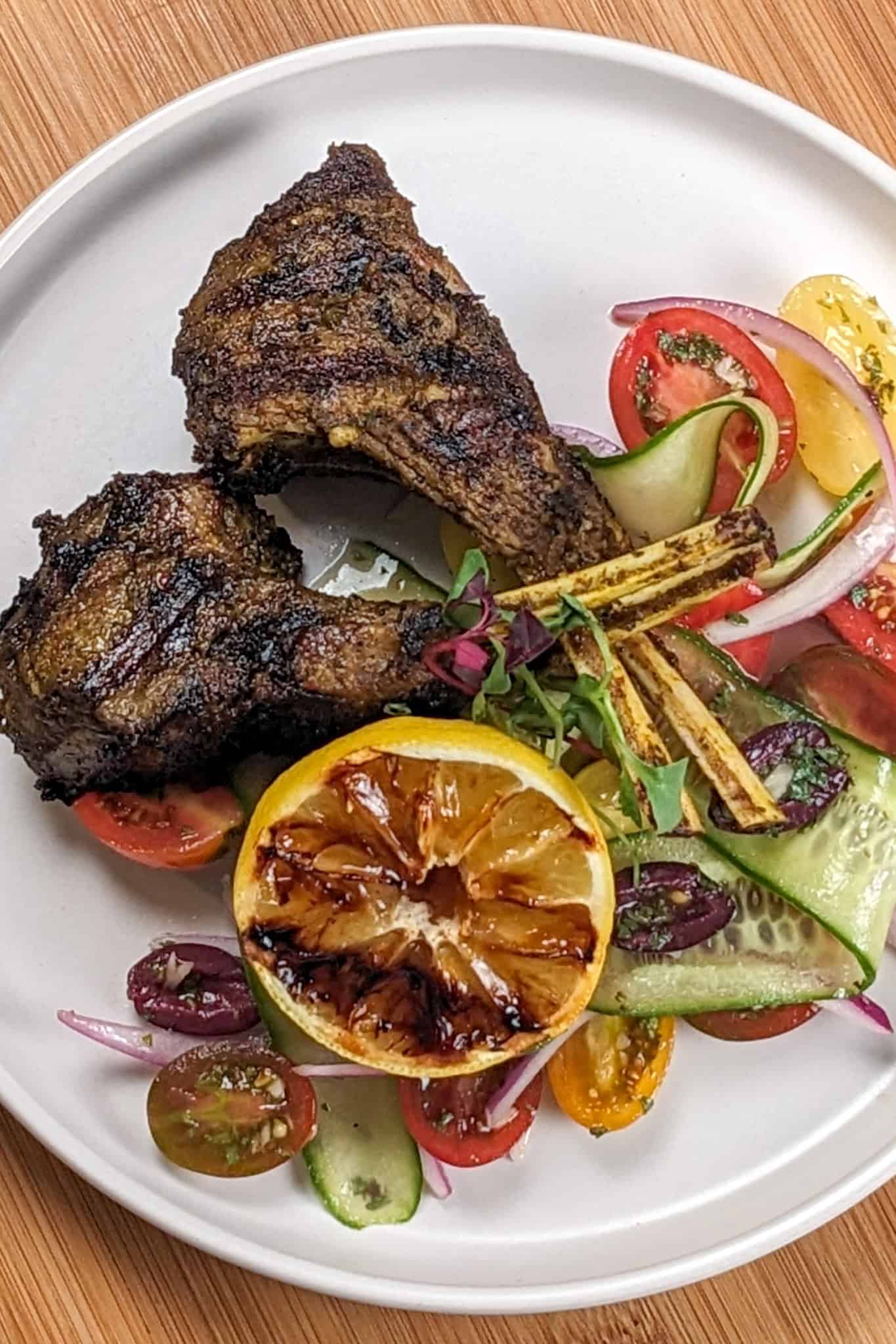 Grilled Zhug Ras el Hanout Lamb Chops and Tomato, cucumber, onion Salad served with a lemon-oregano dressing served with a grilled lemon.