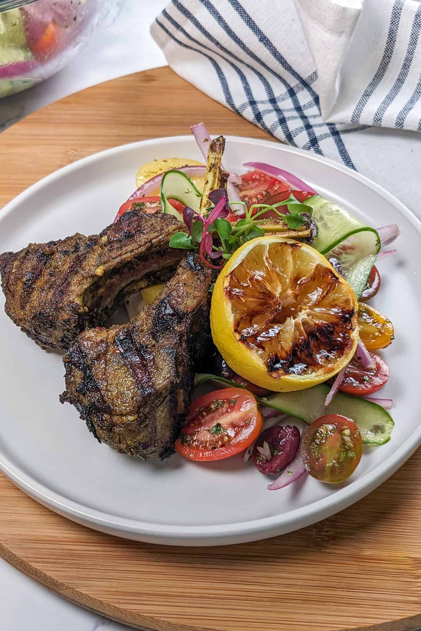 Grilled Zhug Ras el Hanout Lamb Chops and Tomato, cucumber, onion Salad served with a lemon-oregano dressing served with a grilled lemon.