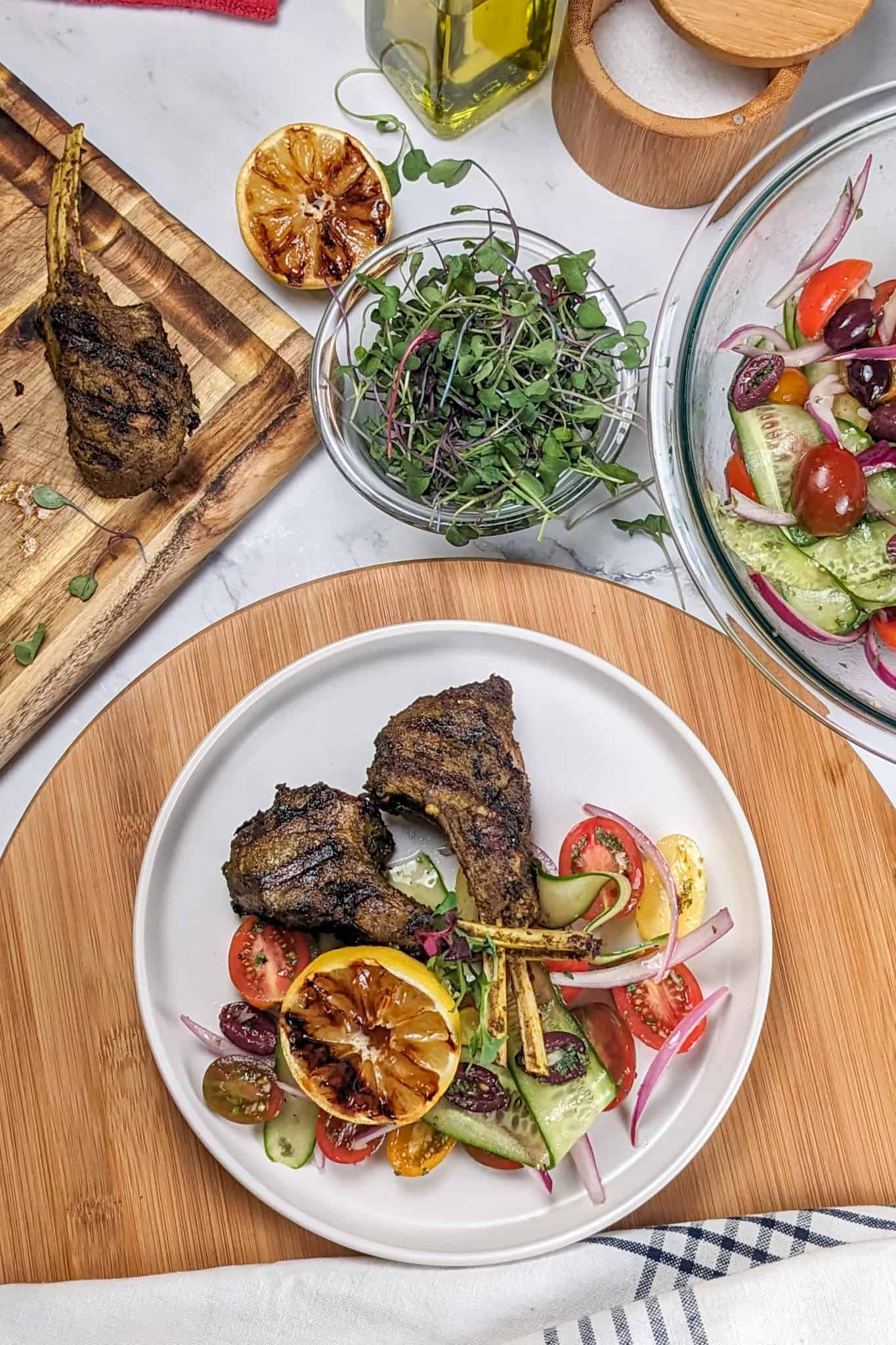 Grilled Zhug Ras el Hanout Lamb Chops and Tomato, cucumber, onion Salad served with a lemon-oregano dressing served with a grilled lemon on a flat plate with shallow straight sides next to more lamb chops on a cutting board, next to a dish of microgreens, and more salad in a mixing bowl.