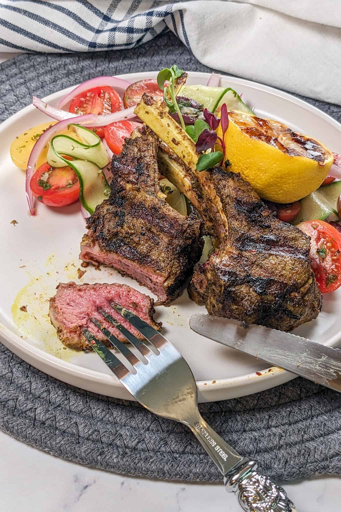 Grilled Zhug Ras el Hanout Lamb Chops and Tomato, cucumber, onion Salad served with a lemon-oregano dressing served with a grilled lemon being eaten.