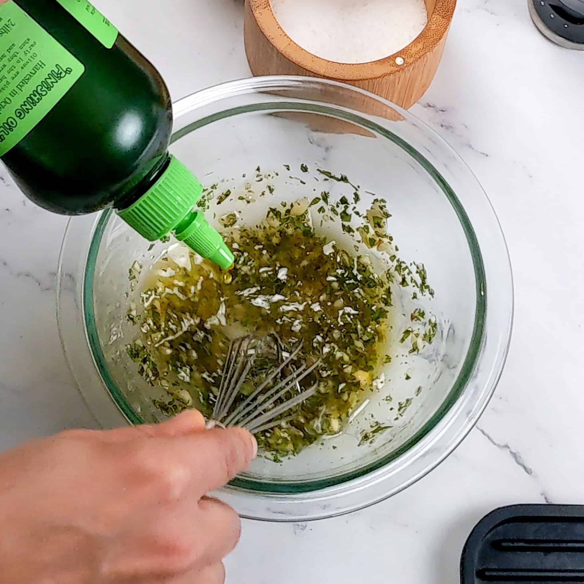 graza extra virgin olive oil being drizzled in while anptjer hand is whisking to make the vinaigrette in a glass mixing bowl.