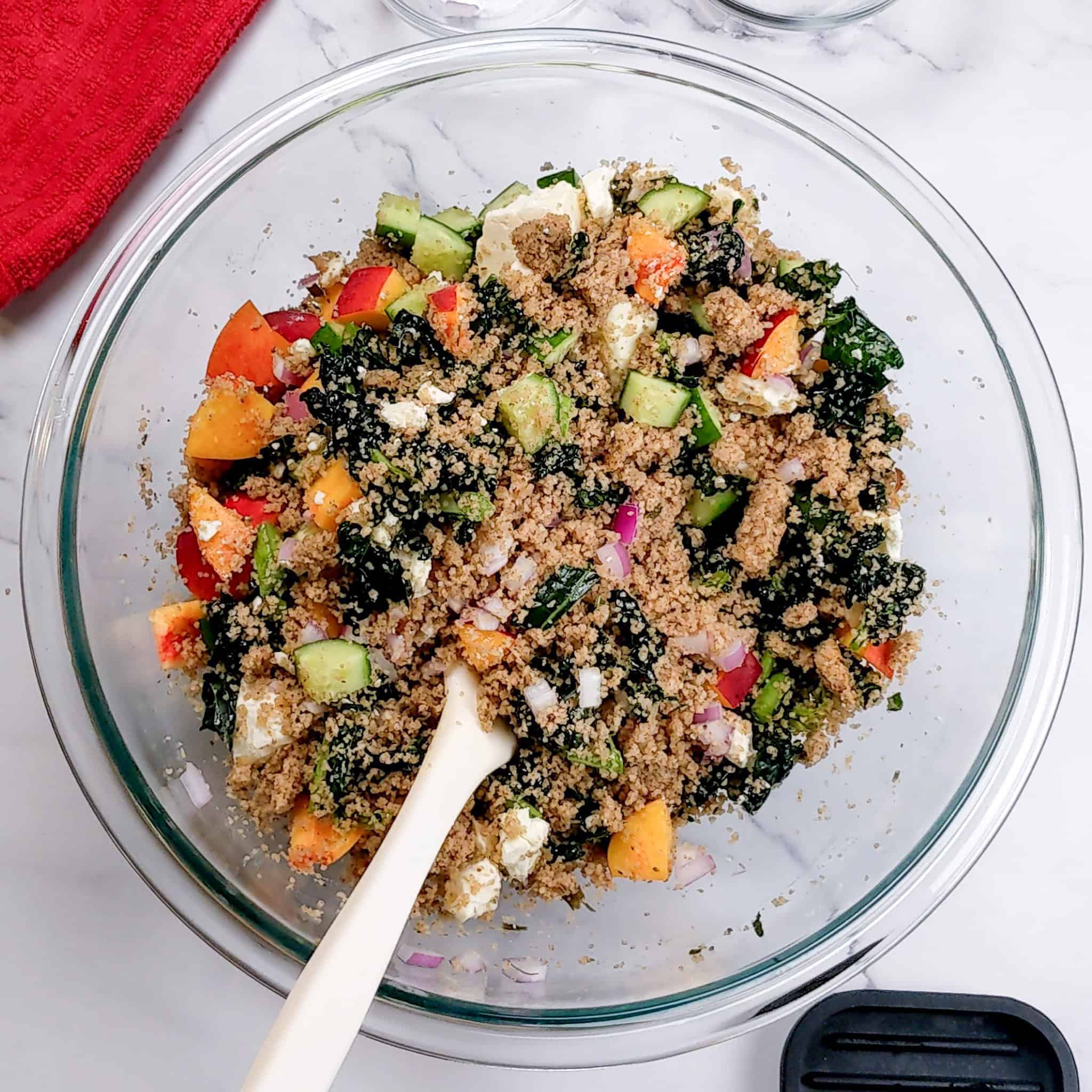 Peach, cucumber, kale, fonio salad with feta cheese tossed in a glass mixing bowl.