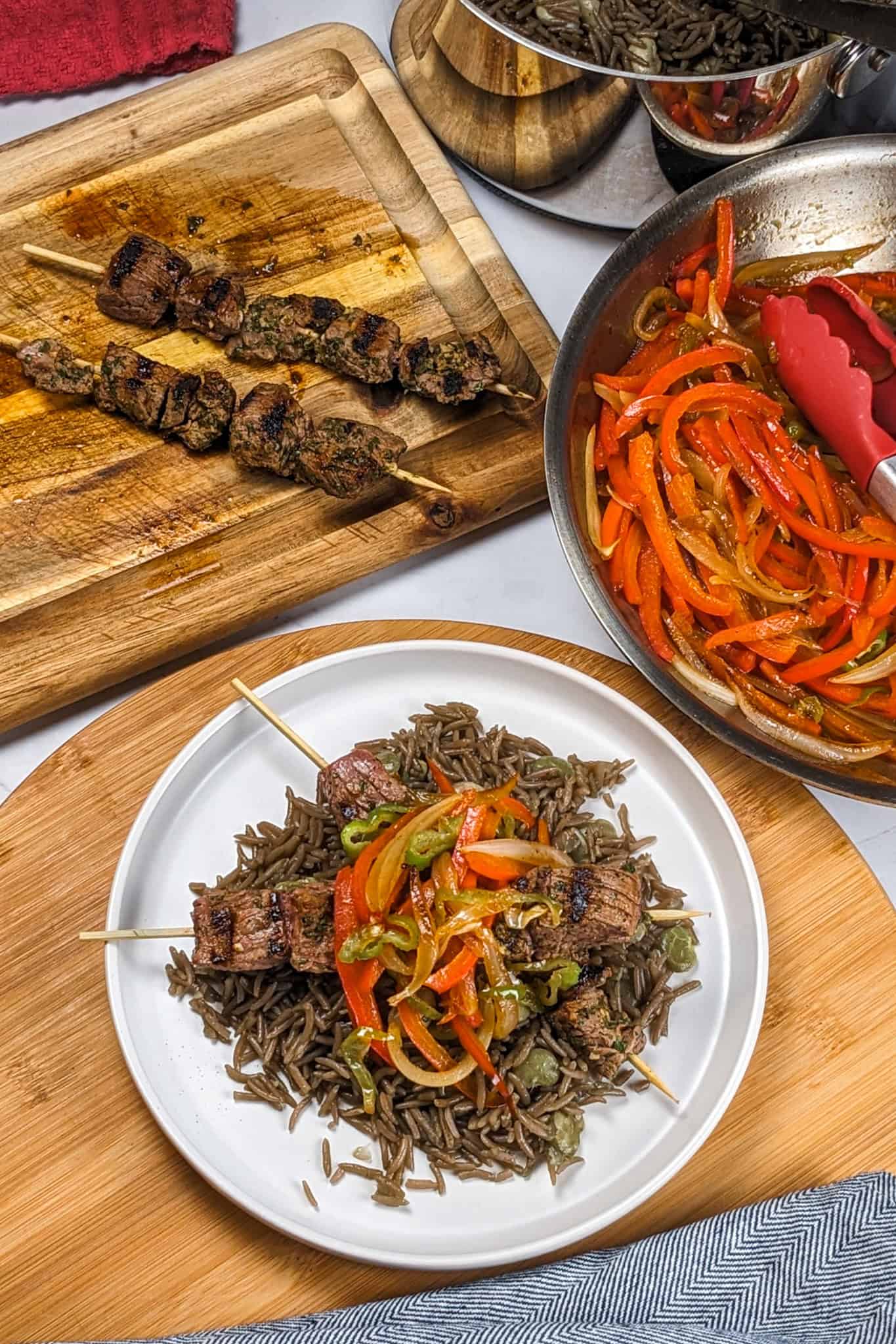 Grilled beef skewers with black mushroom rice on a flat round plate on a wooden lazy Susan next to a kitchen towel, more beef skewers on a cutting board, more cooked rice in a pot, and more escovitch-style vegetables in a saute pan with silicone-tipped tongues.