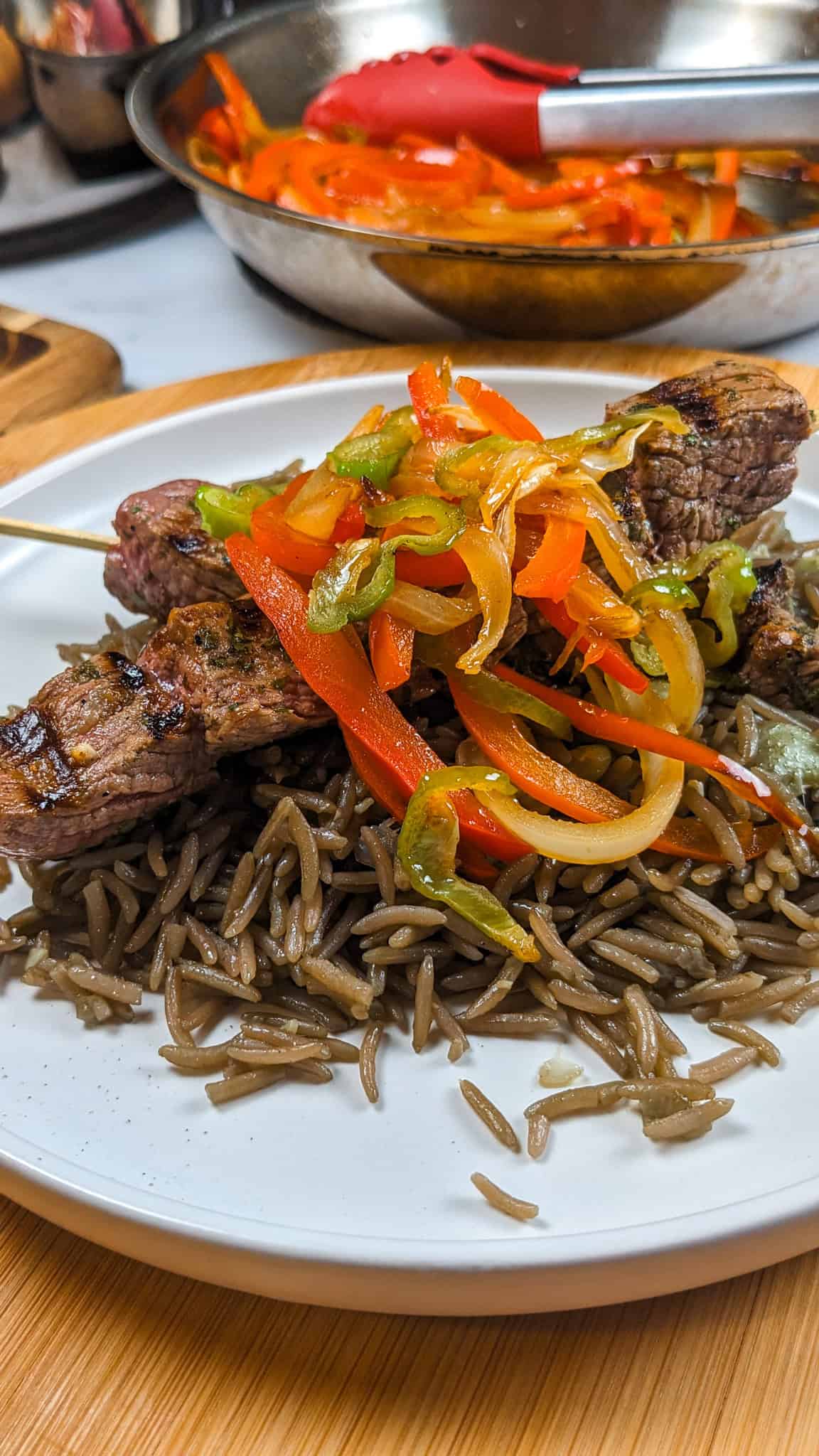 side view of the Grilled beef skewers with black mushroom rice on a flat round plate with more escovitch-style vegetables in the background.