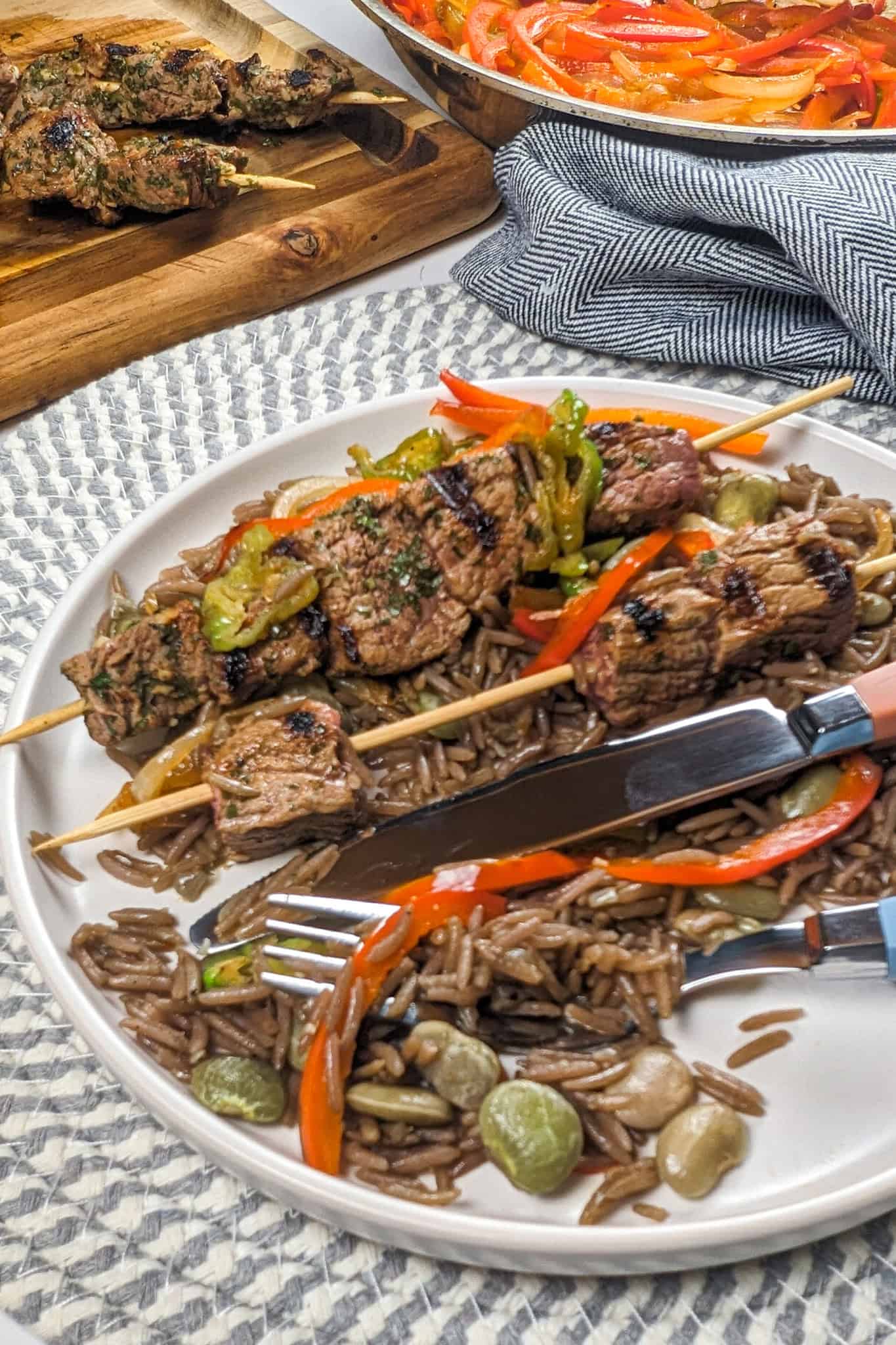 Eaten grilled beef skewers with the black mushroom rice with a knife and fork on the flat round plate that is sitting on a run place mat surrounded by more beef skewers on a cutting board a kitchen towel and more escovitch-style vegetables in a saute pan.