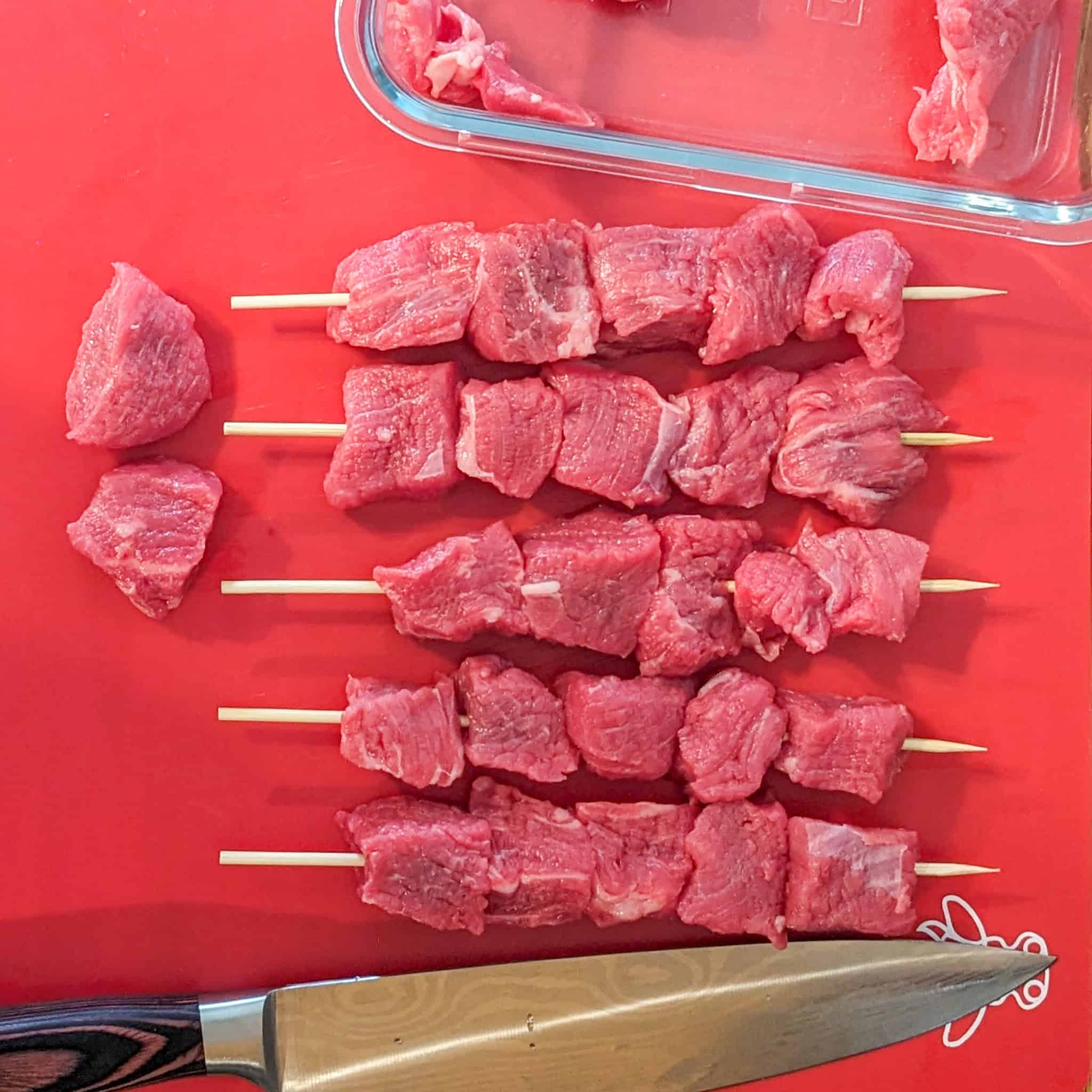 Raw beef chunks on skewers lined up in a row between a rectangle shallow dish with leftover beef chunks and a carving knife.