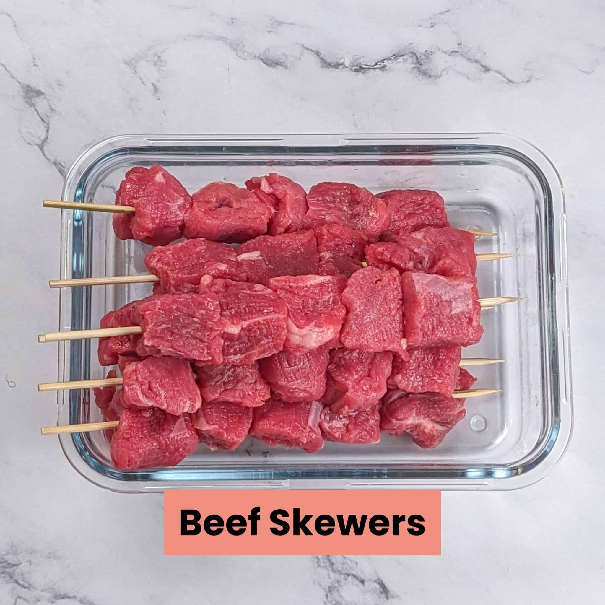 Raw beef chunks on skewers in a rectangular glass dish.