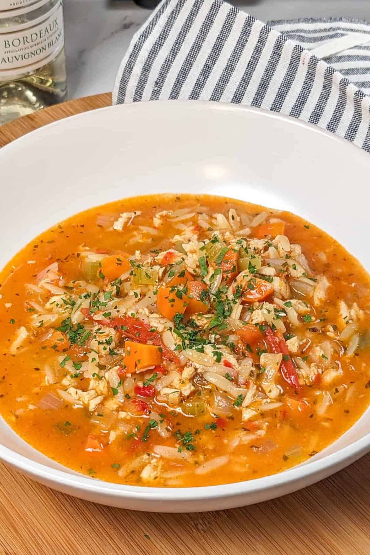 Lemon Calabrian Chili Chicken Orzo Soup in a wide rim bowl on a lazy susan with a kitchen towel and white wine bottle in the background.