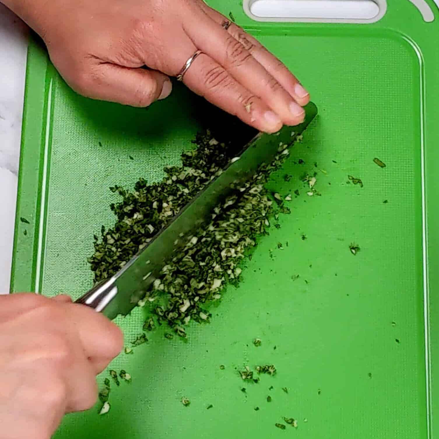 a knife finely chopping garlic and herbs on a plastic cutting board