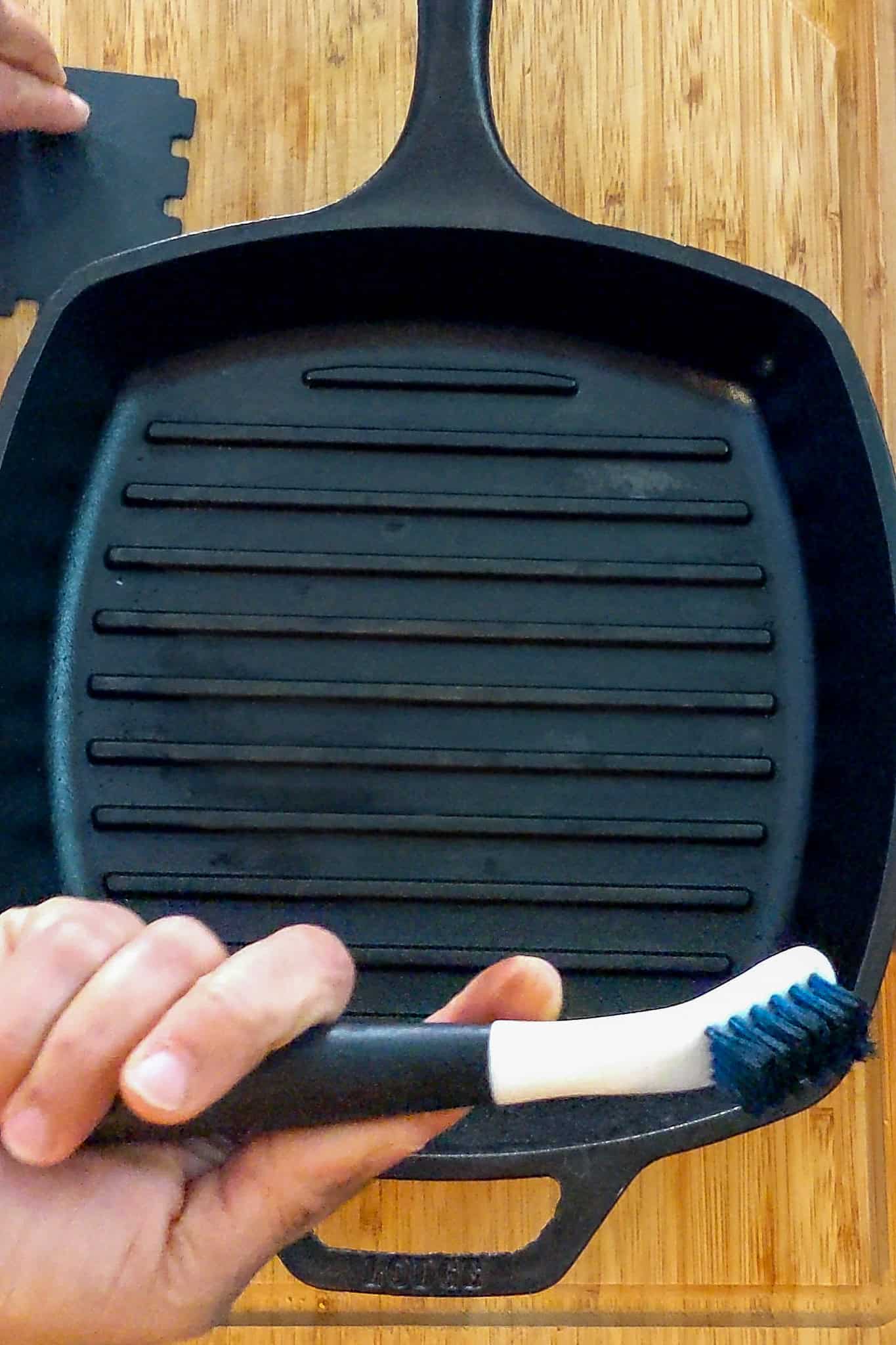 sturdy brush for cleaning cast iron held in hand above cast iron grill pan.