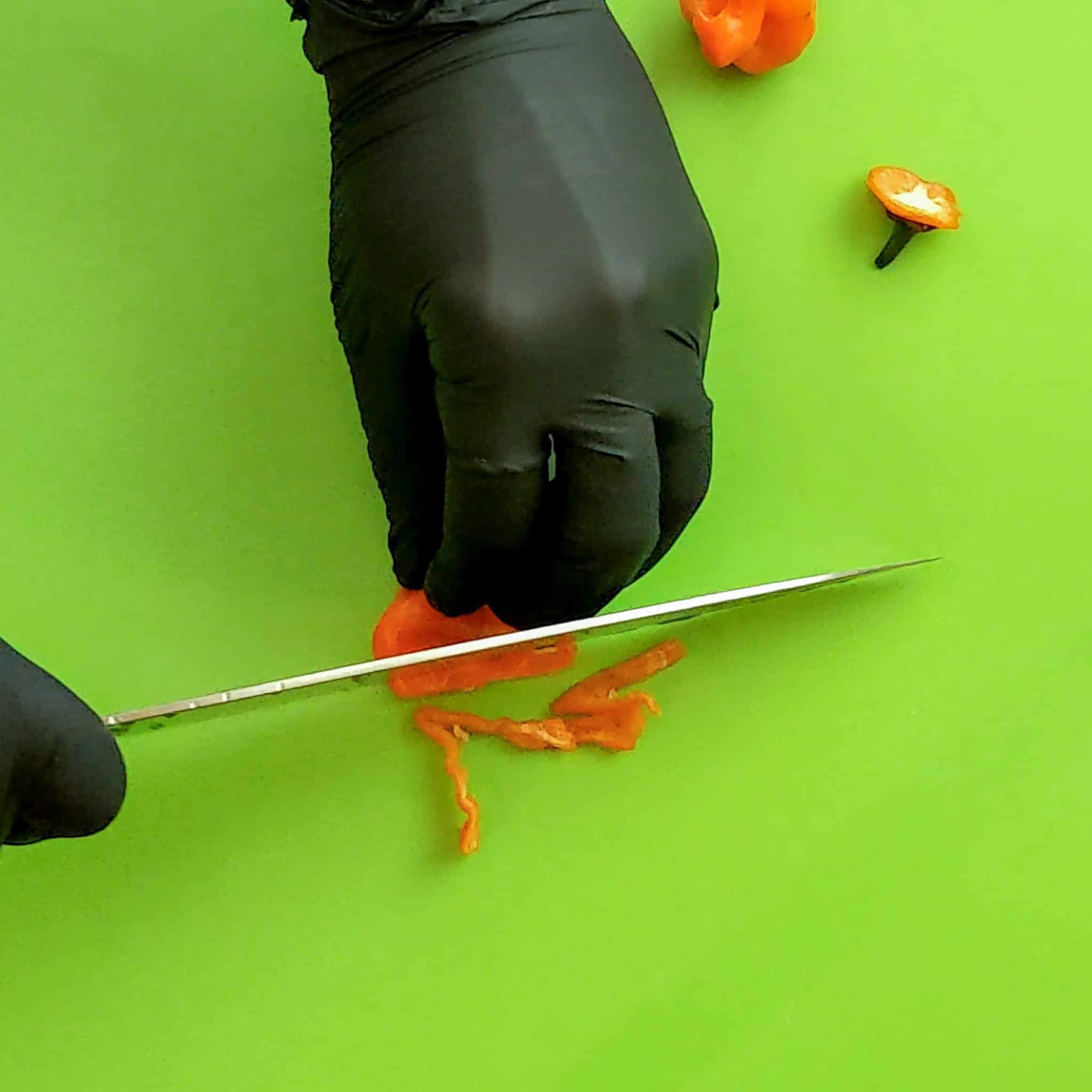 The habanero is cut into thin strips on a color-coded cutting board mat with a sharp knife and gloved hands.