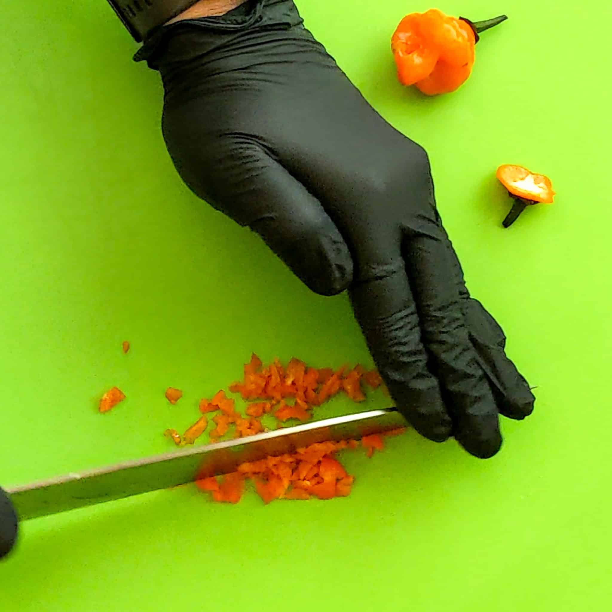 Fresh habanero on a color coded cutting board being chopped with a sharp knife and gloved hand.