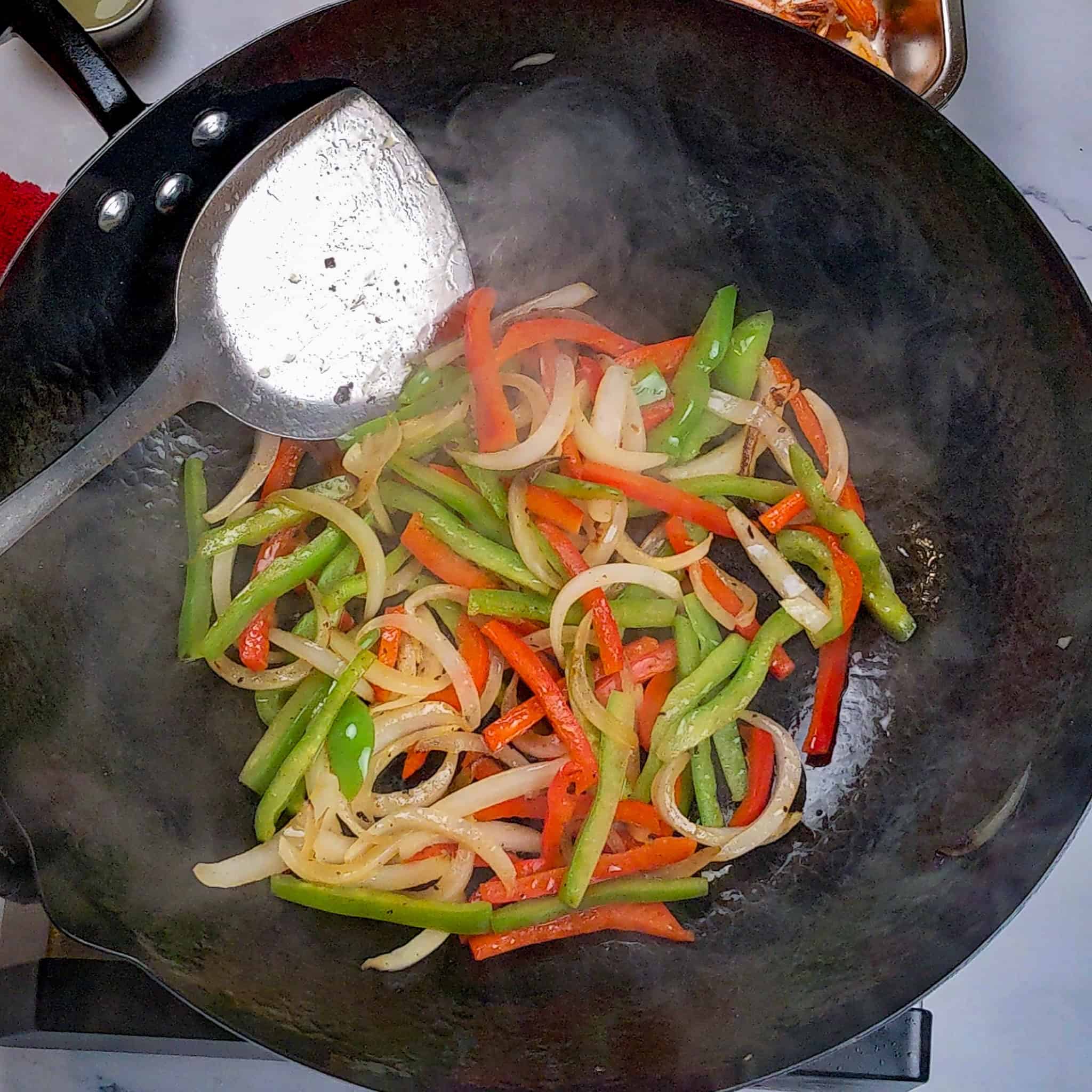 Thinly sliced red bell peppers, green bell peppers, and onions stir-fried in a wok with a wok spatula.