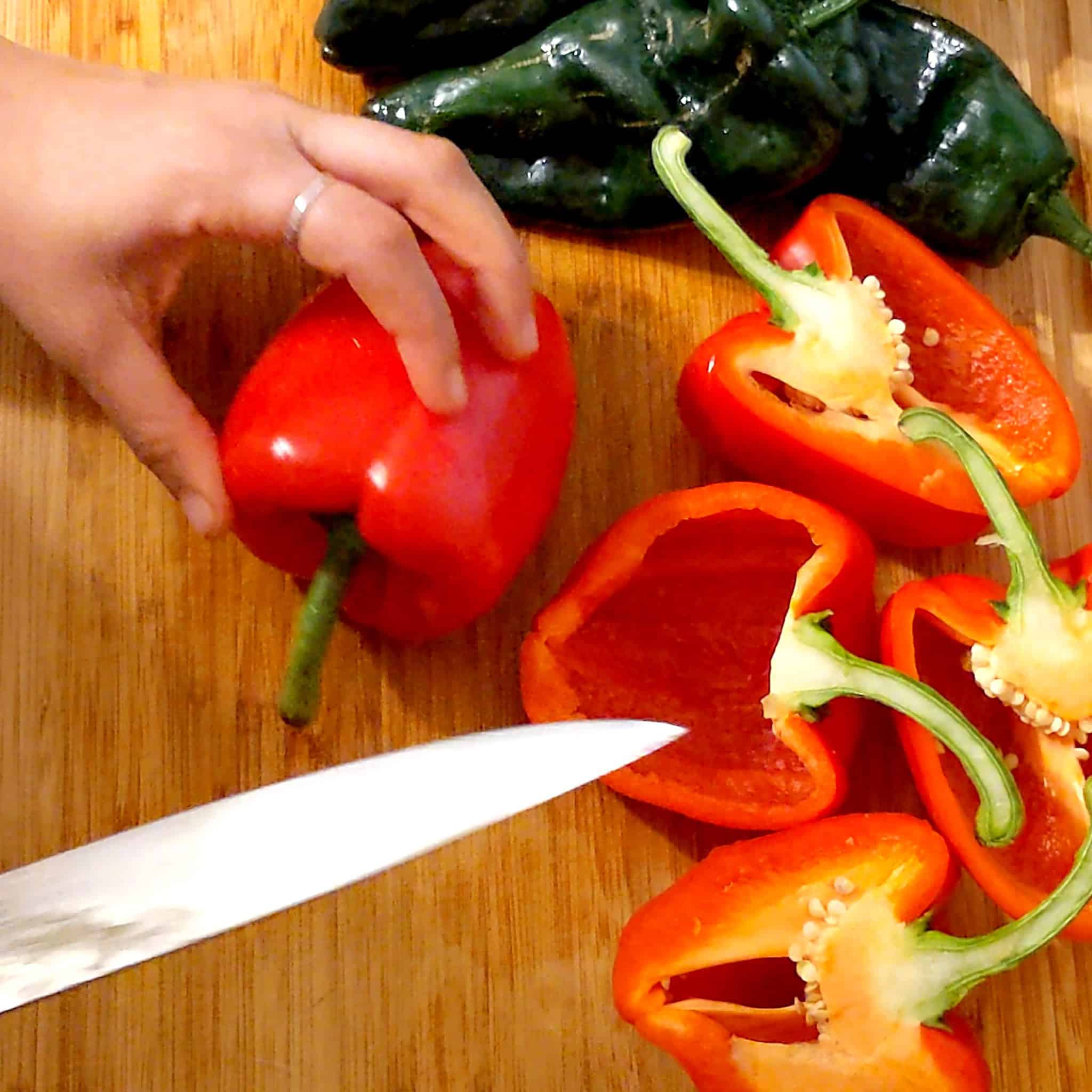 a red bell pepper on its side surrounded by halved peppers and poblano peppers with a knife.