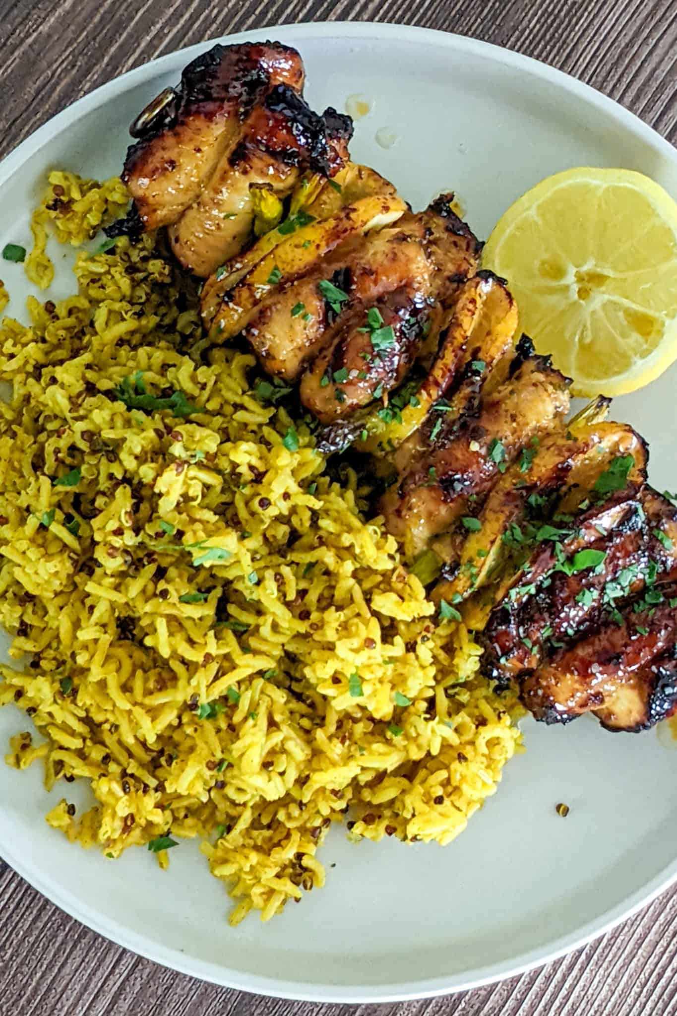 slightly charred cooked hot Honey lemon glazed chicken skewers served with a side of yellow rice and a half of a lemon on a round flat plate.