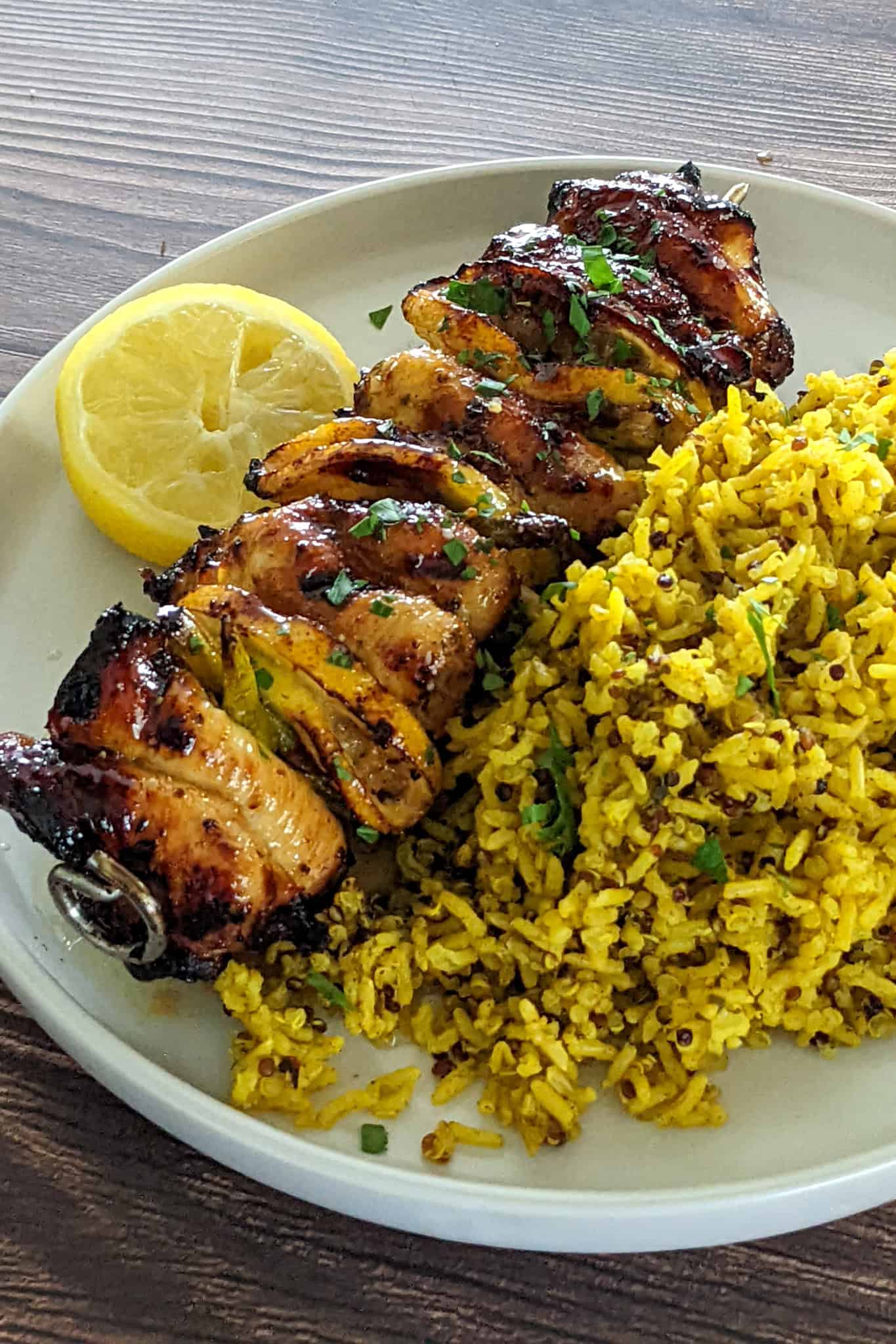 slightly charred cooked hot Honey lemon glazed chicken skewers served with a side of yellow rice and a half of a lemon on a round flat plate.