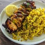 slightly charred cooked hot Honey lemon glazed chicken skewers served with a side of yellow rice and a half of a lemon on a round flat plate.