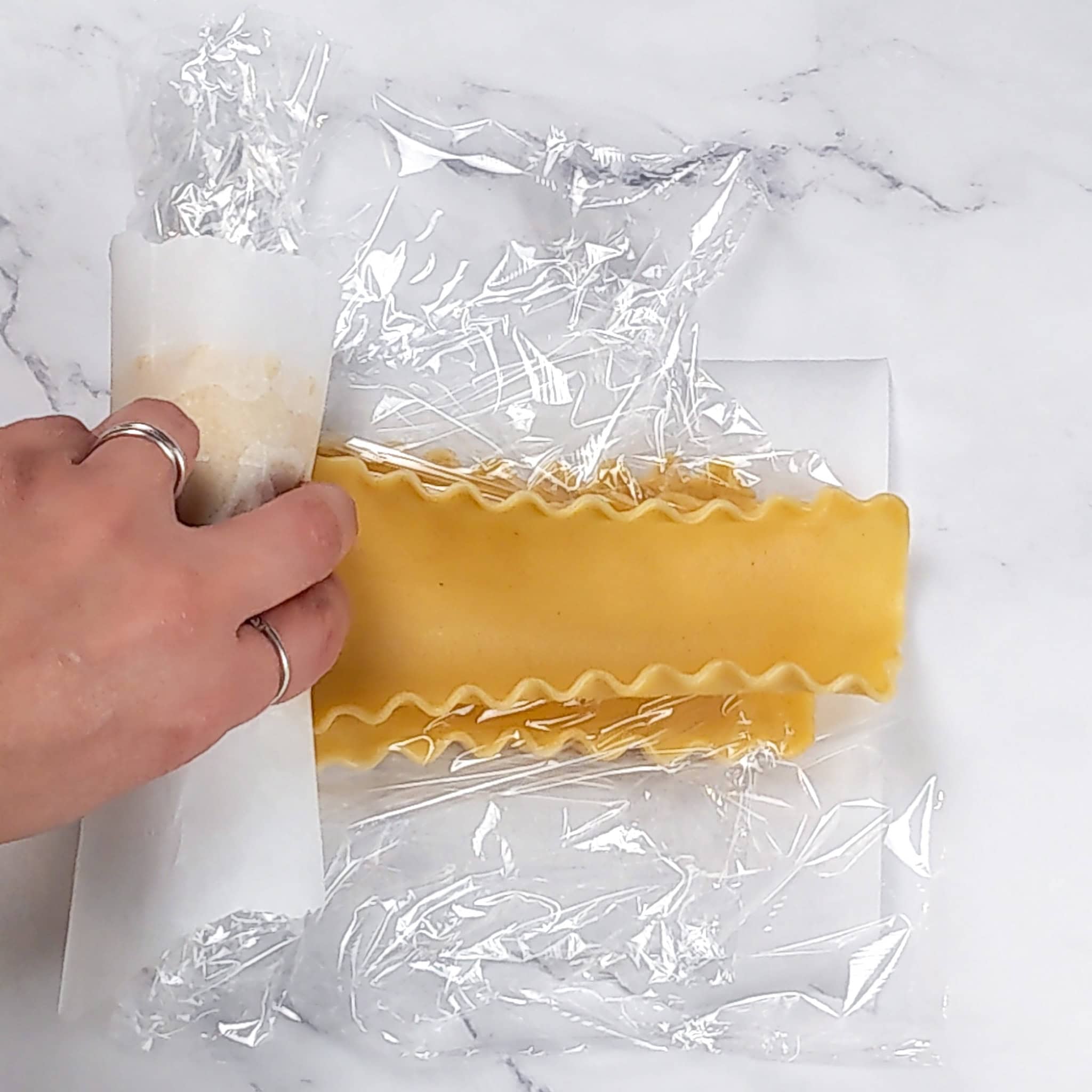 parchment paper layered lasagna sheets being rolled with plastic film wrap.