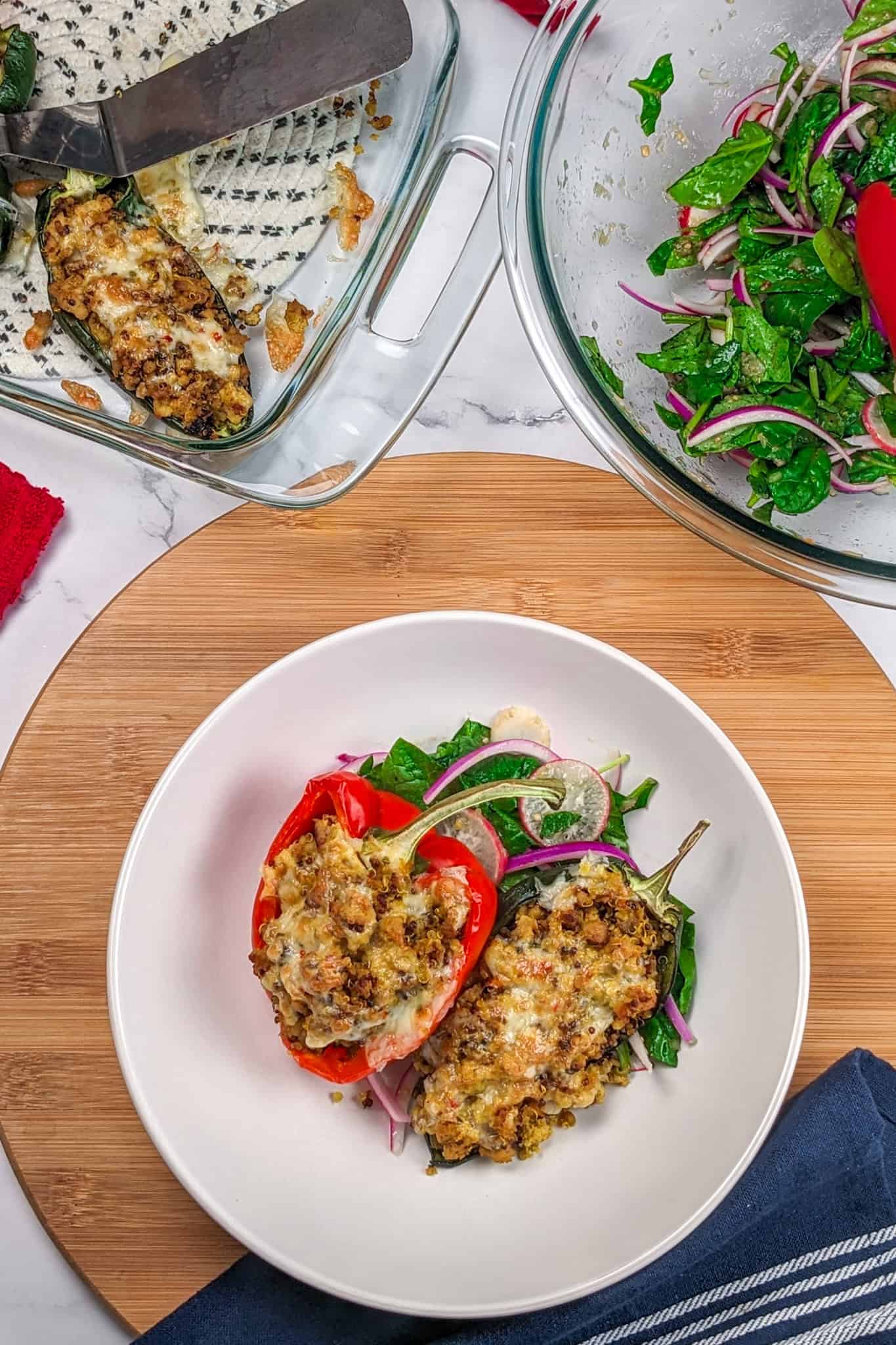 Baked stuffed bell peppers and poblano peppers with ground turkey and quinoa with melted pepper jack cheese on a bed of greens with sliced radishes and red onion. Surrounded by a baking dish of leftover stuffed peppers and a mixing bowl of salad.