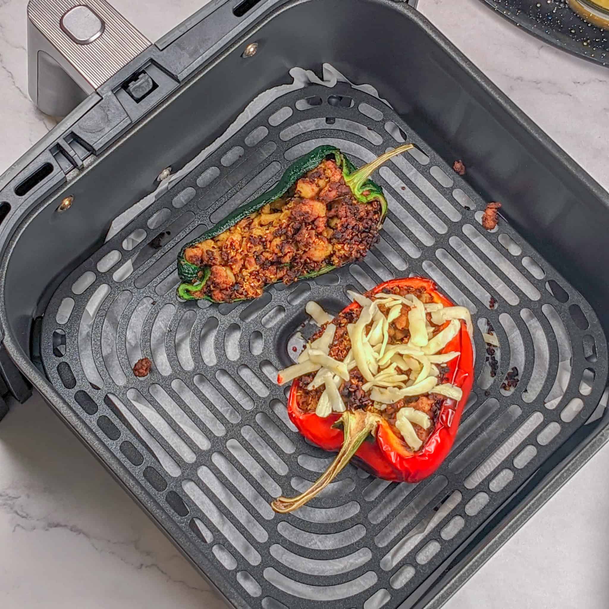 Stuffed bell pepper and poblano pepper laying in an air fryer basket with shredded cheese on the red bell pepper.