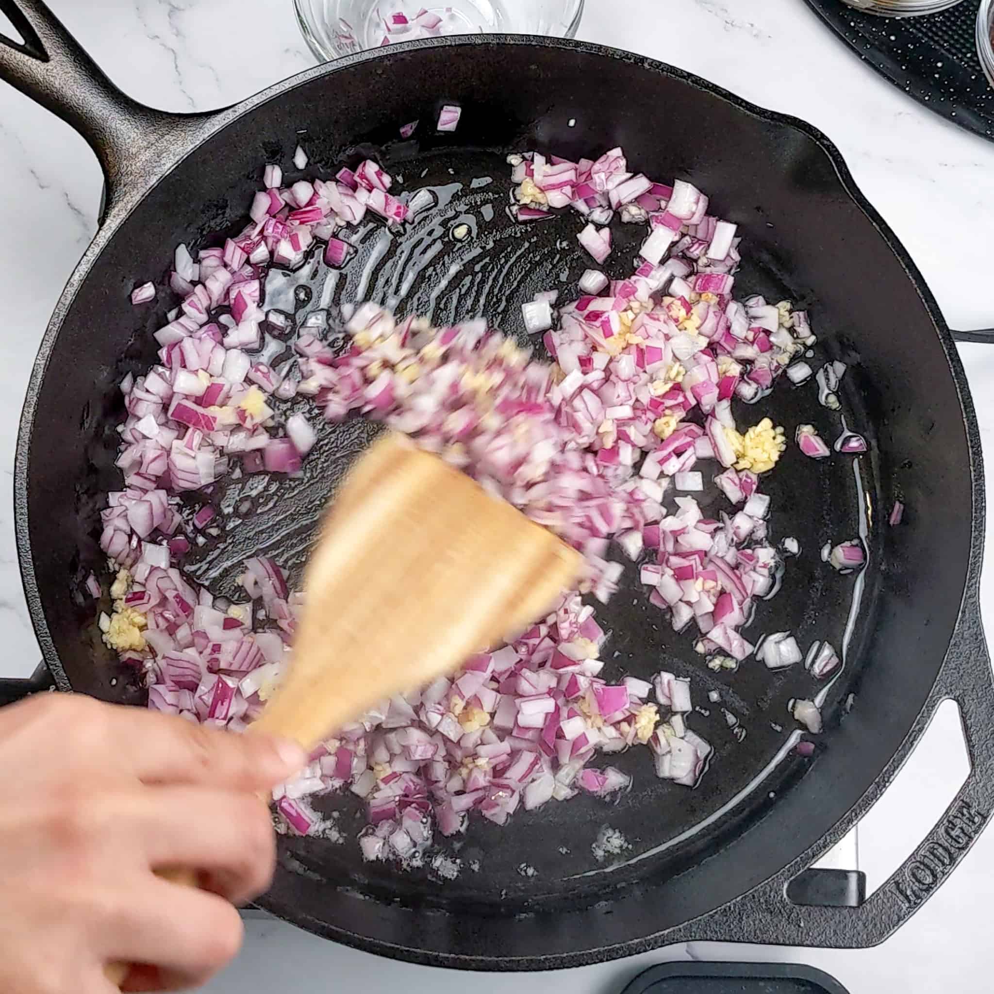 Chopped red onion and garlic sauteing in a cast iron skillet and stirred with a wooden spatula.