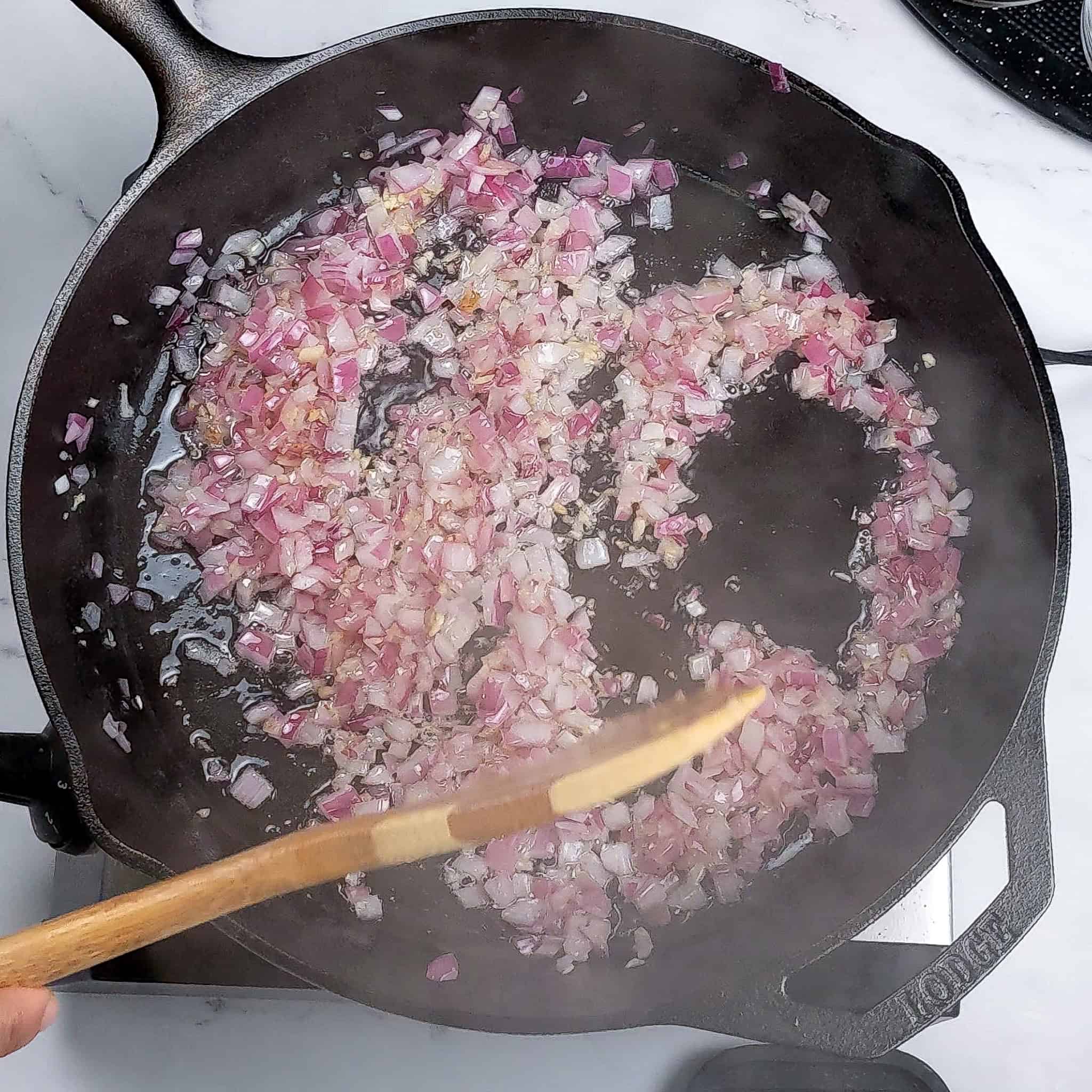 Cooked chopped red onion and garlic sauteing in a cast iron skillet and stirred with a wooden spatula.