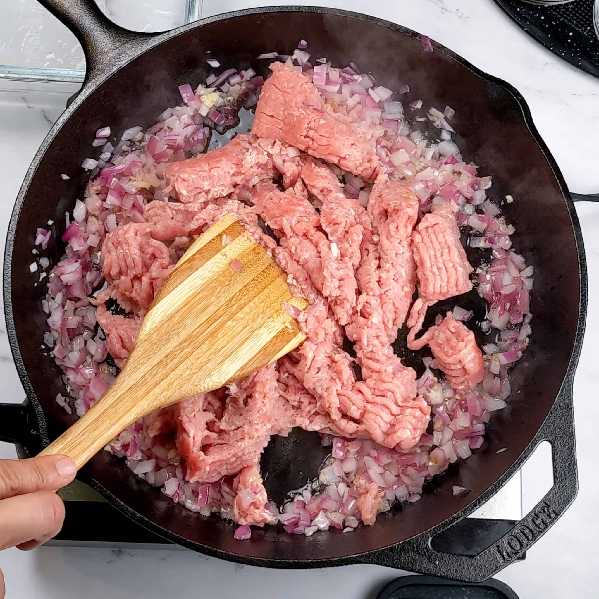 Raw ground turkey is chopped up with a wooden spoon and mixed with the cooked chopped onion and garlic.