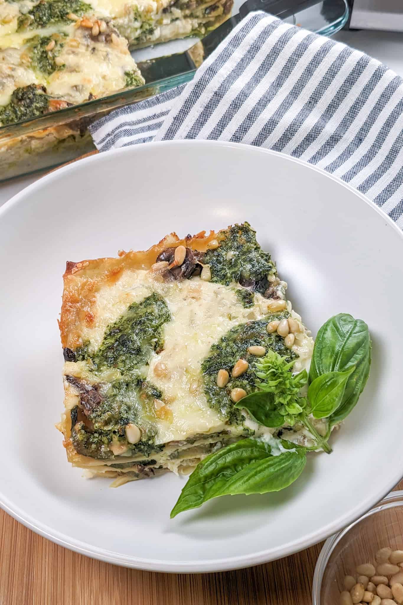 Creamy Spinach Pesto Mushroom Lasagna on a wide rim pasta bowl garnished with a fresh basil leaves surrounded by the baking dish, kitchen towel and more pine nuts.