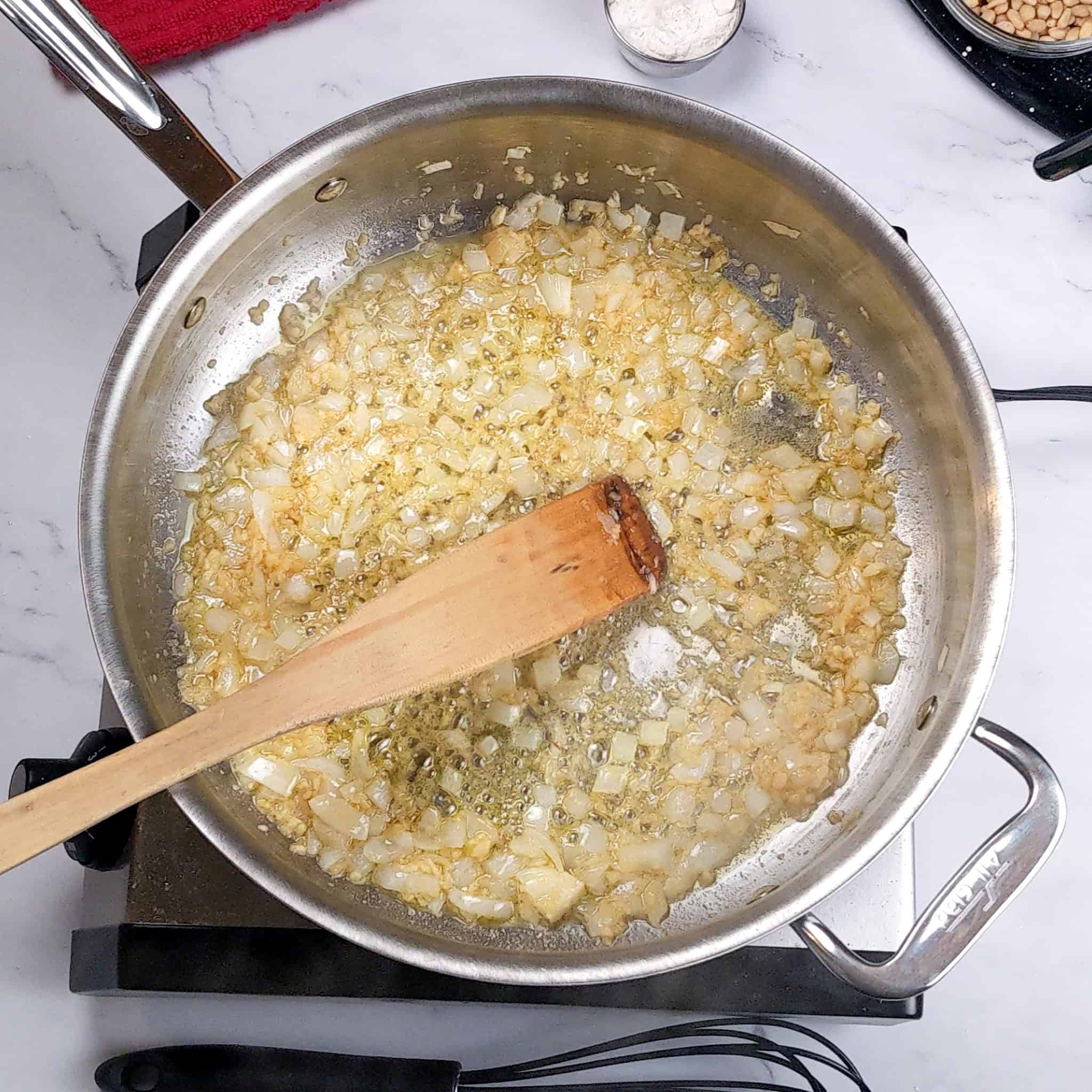 chopped onion and garlic browning in an all clad stainless steel sauce pan being stirred with a wooden spatula.