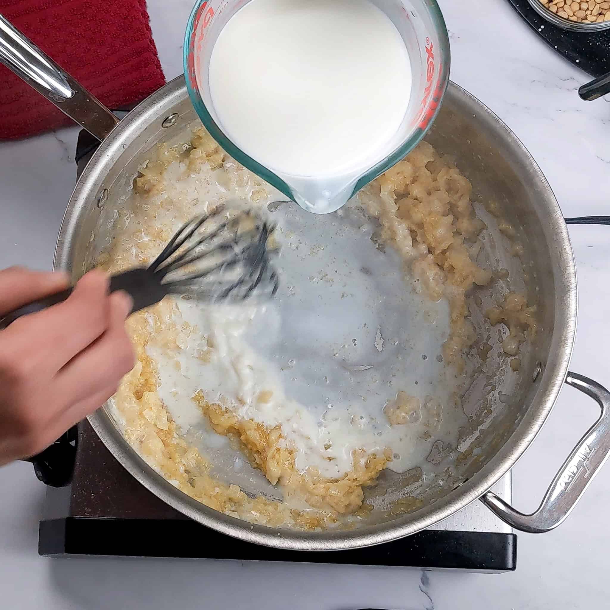 Milk in a Pyrex glass measuring cup is poured while the other hand whisks into the onion and garlic, which has been browned and thickened with flour.