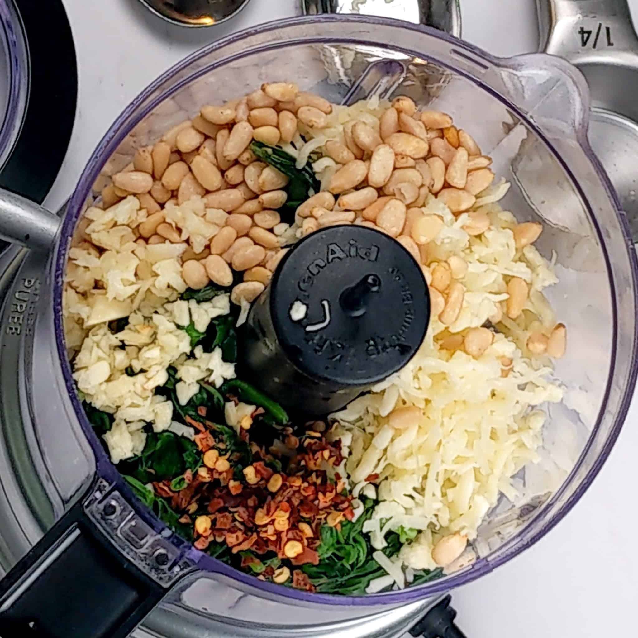 Top view the open food processor with pine nuts, chopped garlic, crushed red pepper flakes, cheese blend, and blanched basil and spinach.