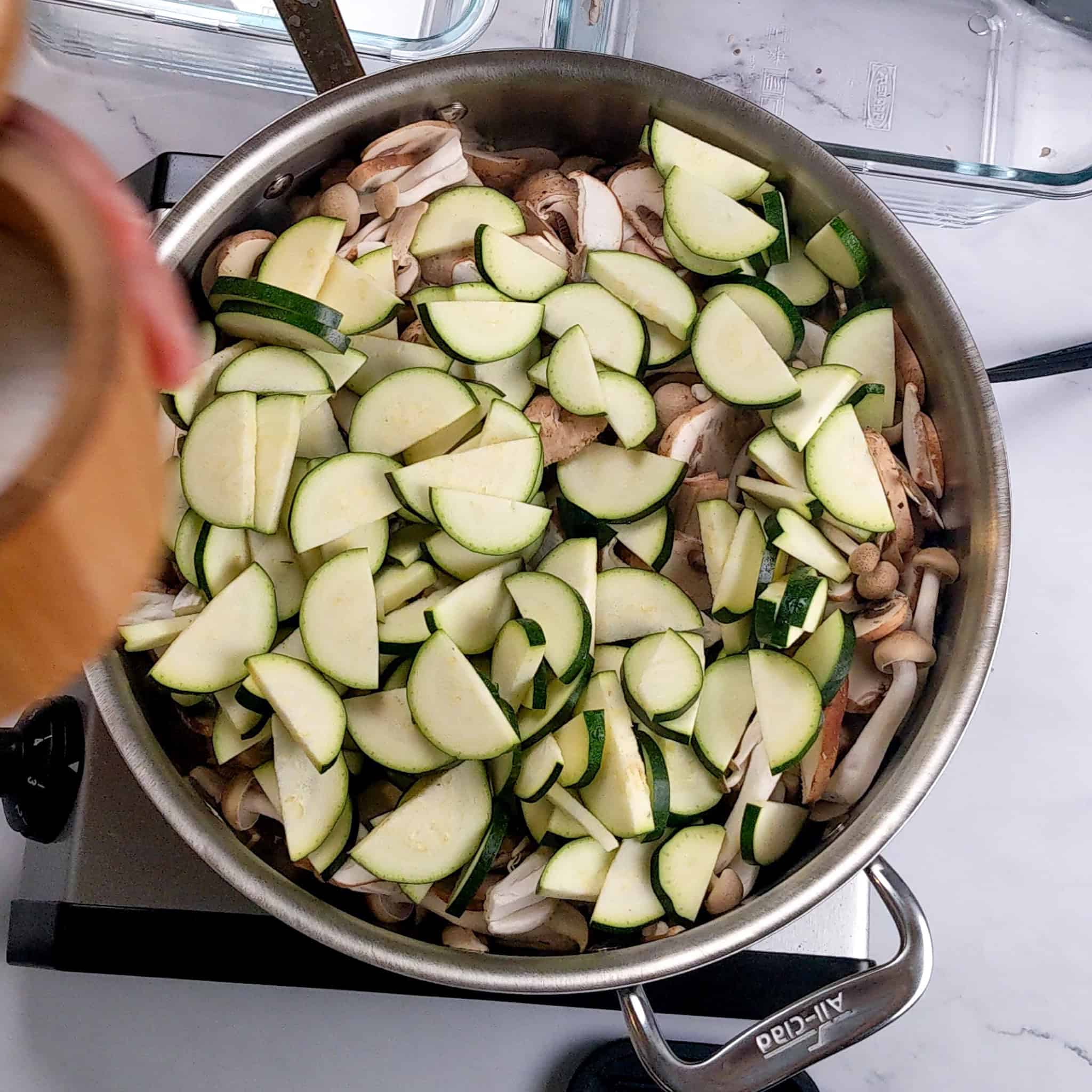 The All- clad saute pan with tall sides made out of stainless steel is filled with sliced mushrooms and zucchini.