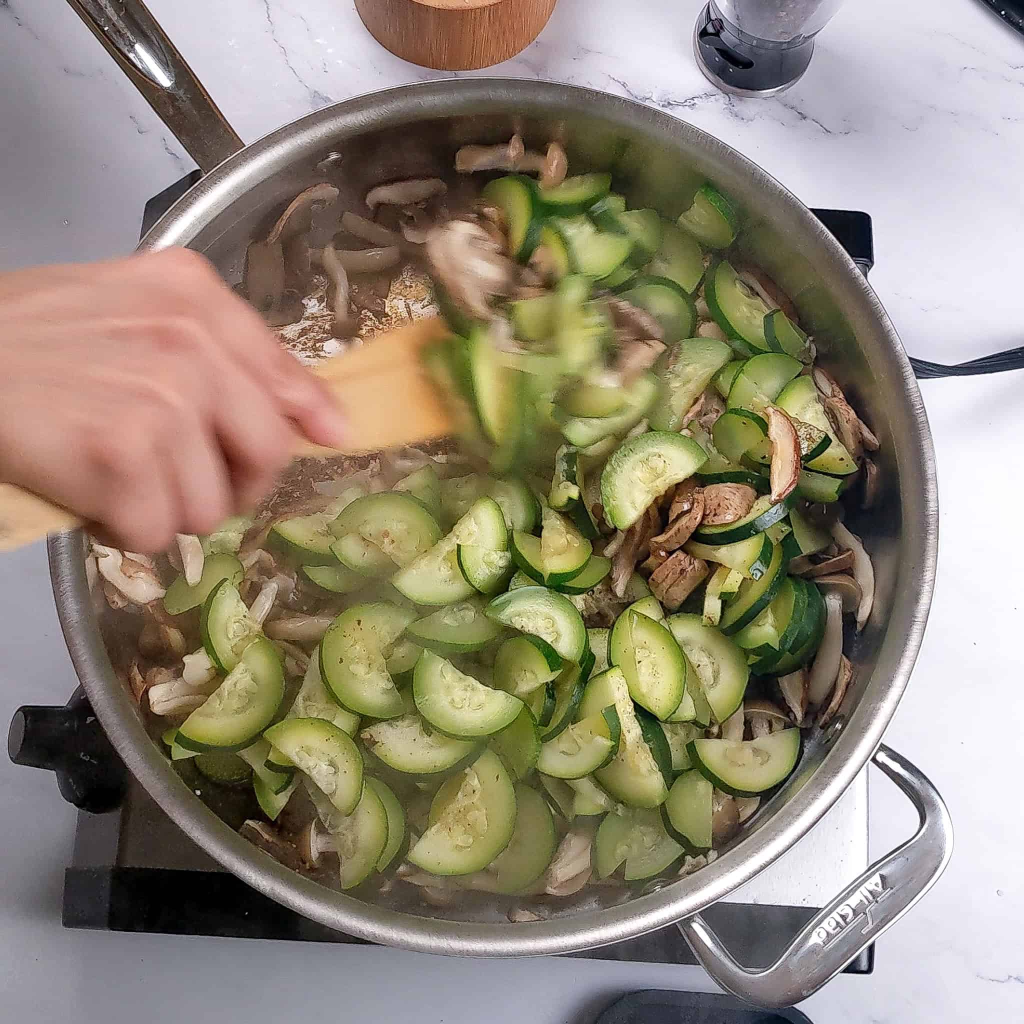 The sliced cooked mushroom and zucchini are stirred with a wooden spatula in the saute pan.