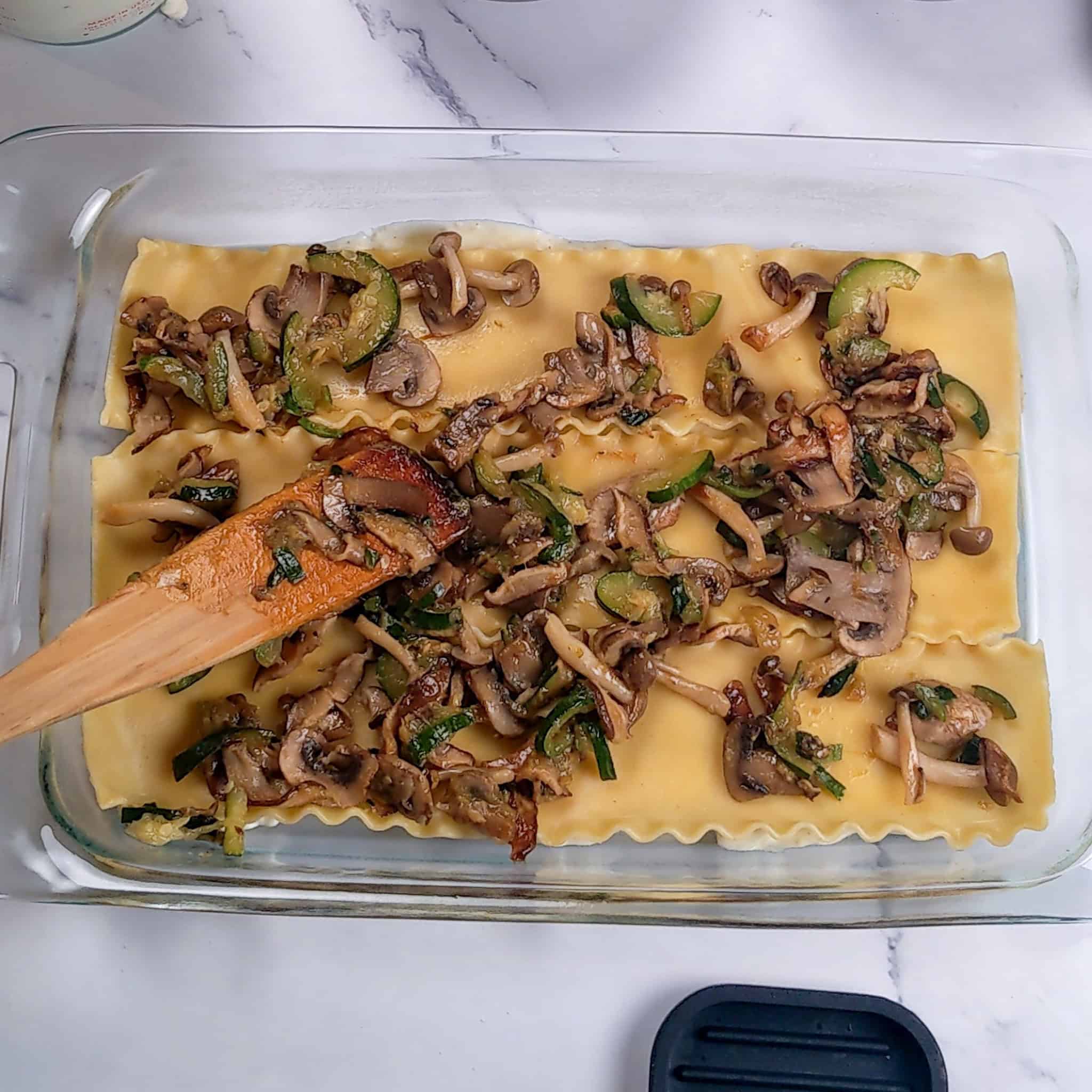 The cooked mushrooms and zucchini layered on top of the lasagna sheets with a wooden spatula in the glass baking dish.