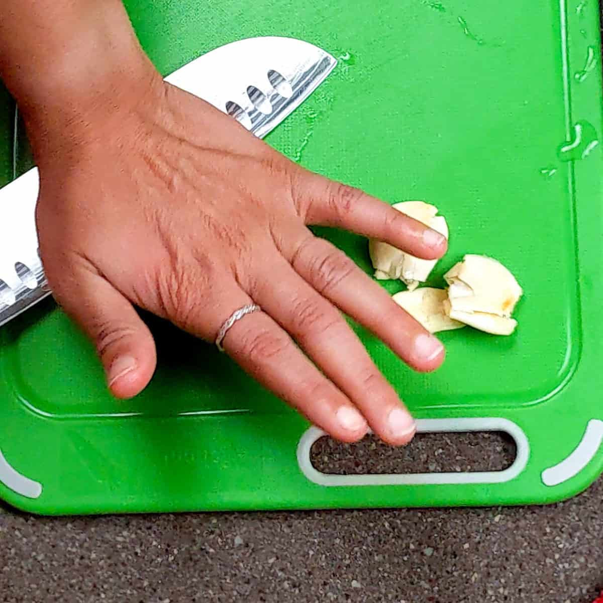 Hand chopping garlic on a vibrant green plastic cutting board with a sharp kitchen knife.