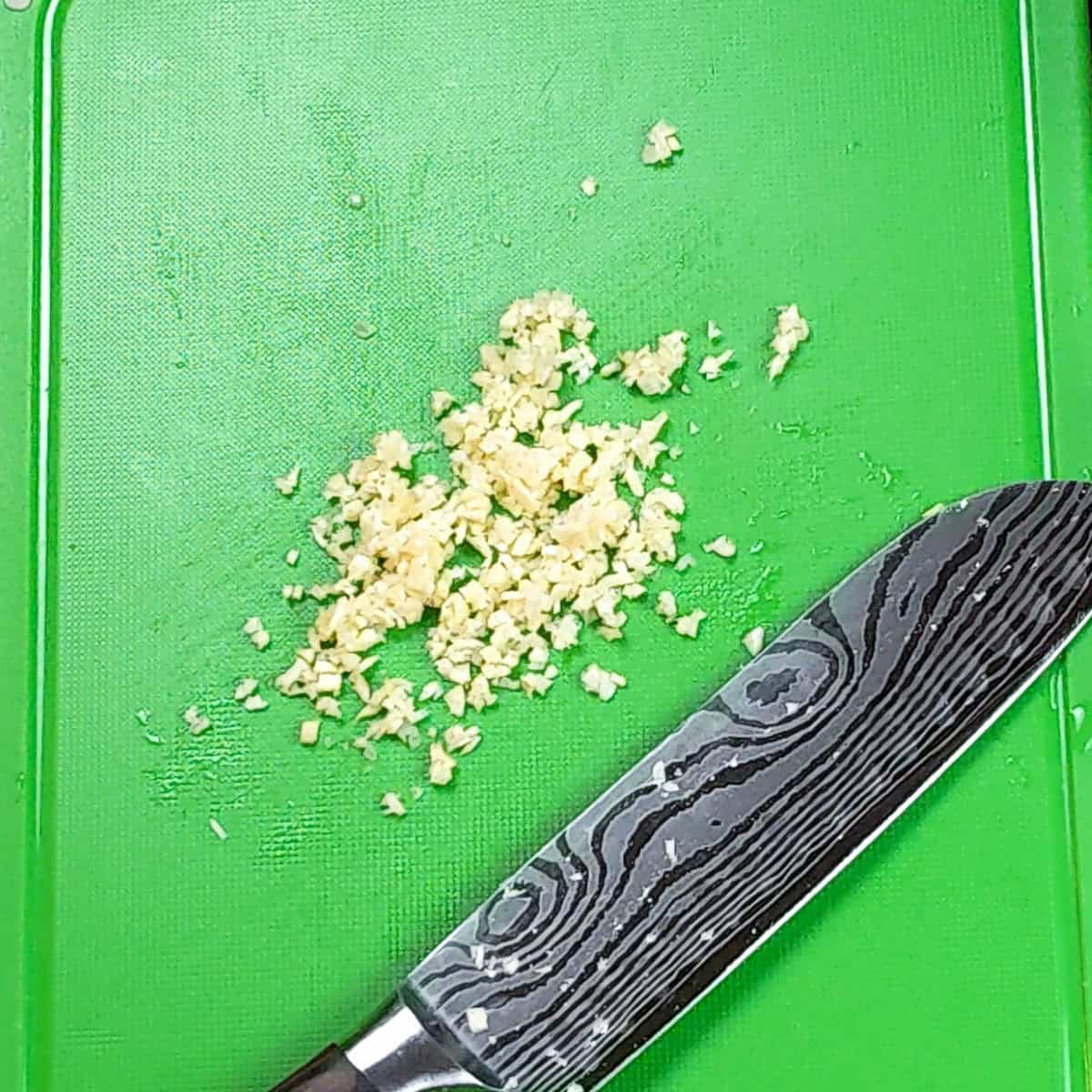 Freshly chopped garlic on a green cutting board with a quality Damascus knife.