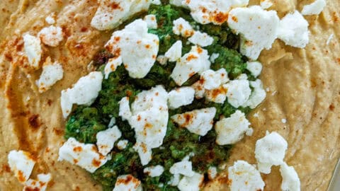 close up of chunky hummus topped with zhug in the center and sprinkled with crumbled feta cheese and smoked paprika and drizzled with olive oil.